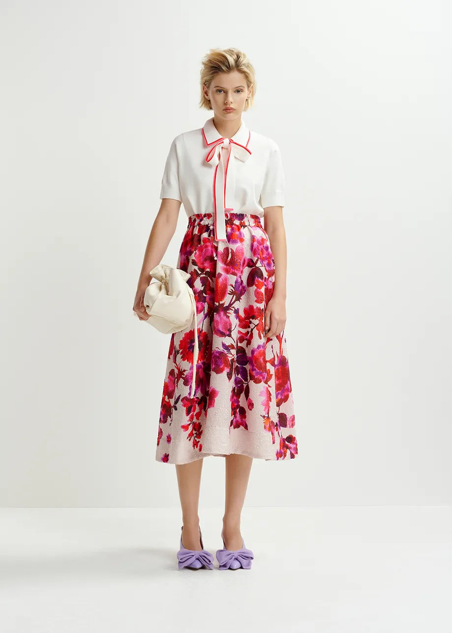 An individual in an Essentiel Antwerp Hight Knitted Polo blouse with ribbed trims and a floral skirt, holding a beige handbag and wearing purple shoes, stands against a plain background.