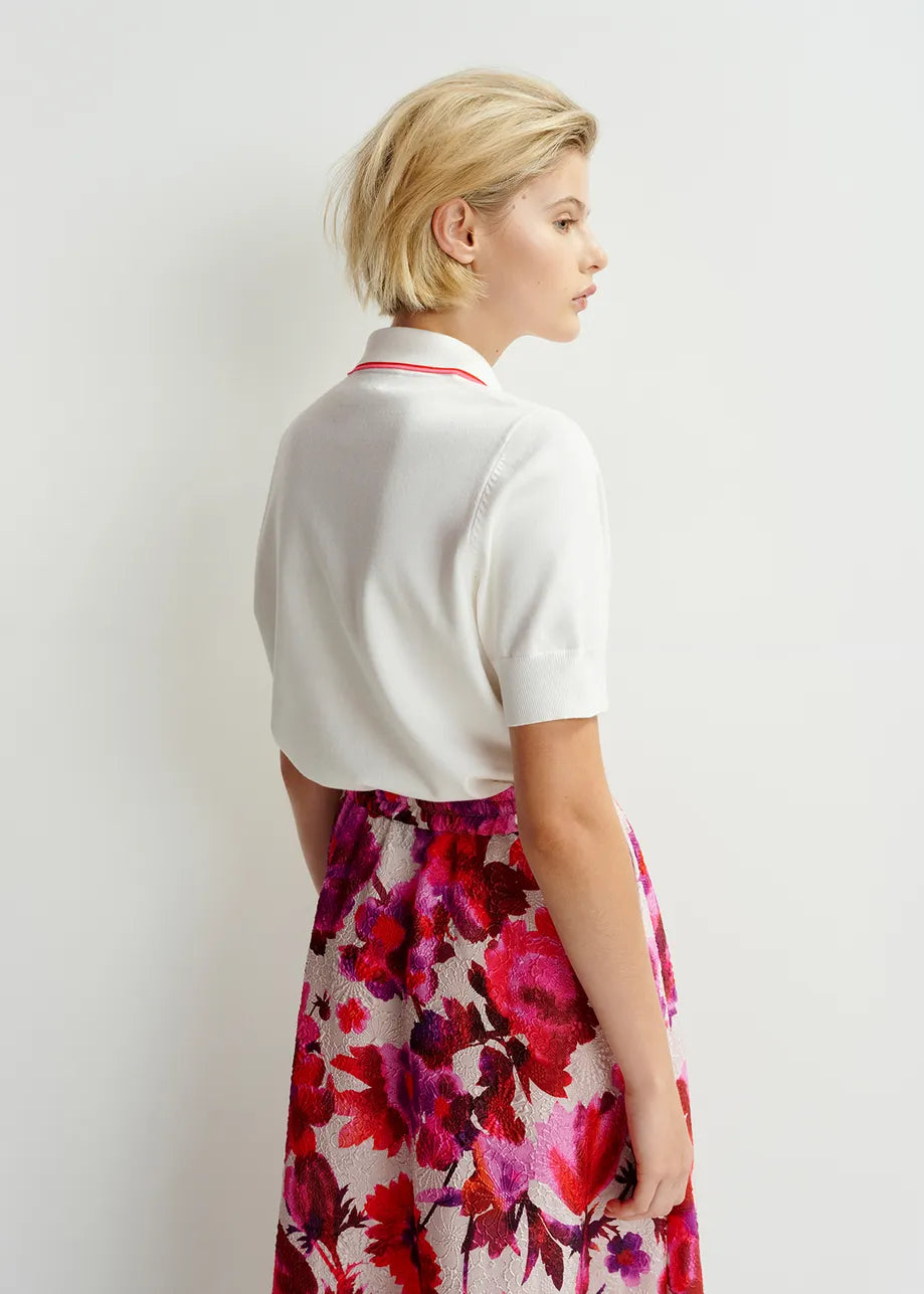 A woman with short blonde hair models the Essentiel Antwerp Hight Knitted Polo, a chic white top with ribbed trims, and pairs it with a pink floral skirt while facing sideways against a plain white background.