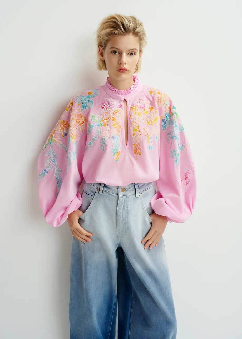 A person wearing the Essentiel Antwerp Hitrus Blouse in pink, featuring floral embroidery and balloon sleeves, paired with wide-leg jeans, is standing against a plain backdrop.