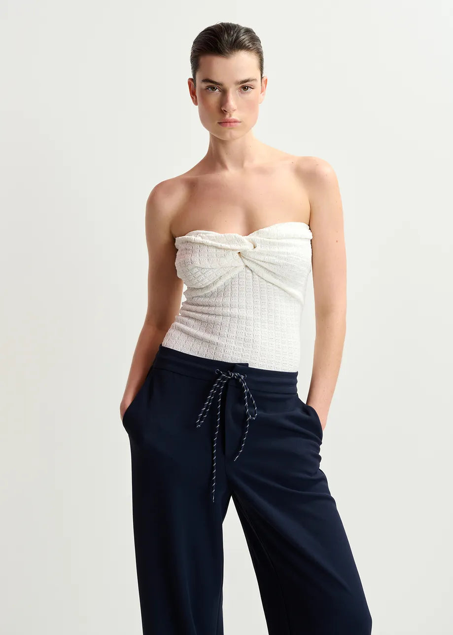 A person wearing an Essentiel Antwerp Holo Knitted Strapless Top in white and navy high-waisted bottoms with hands in pockets stands against a simple backdrop.