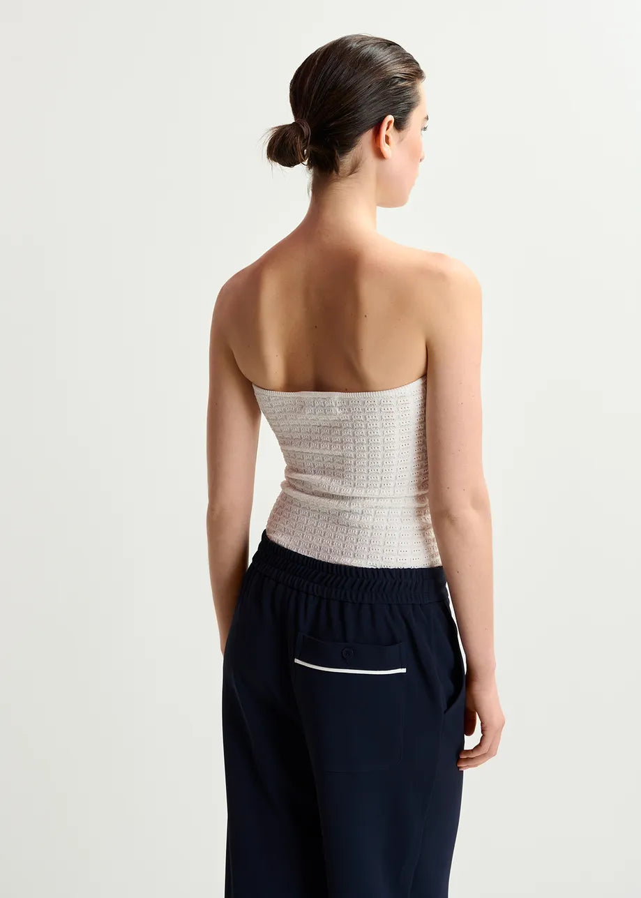 A person with dark hair tied back wears the Essentiel Antwerp Holo Knitted Strapless Top in white and navy high-waisted bottoms, standing against a plain light background.