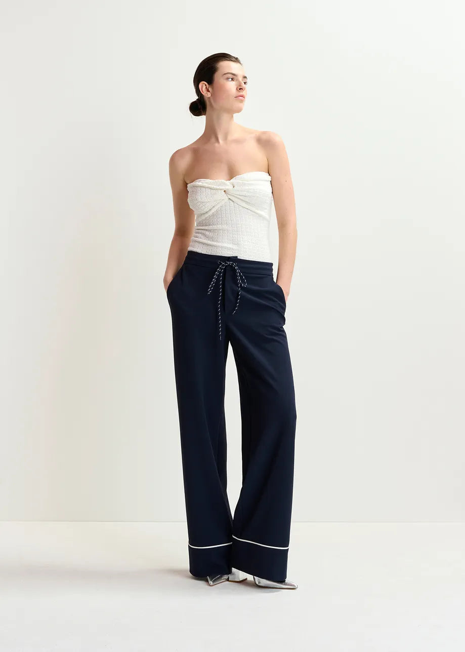 Person in a Holo Knitted Strapless Top - White by Essentiel Antwerp paired with dark high-waisted wide-leg pants, hands casually in pockets against a plain background.