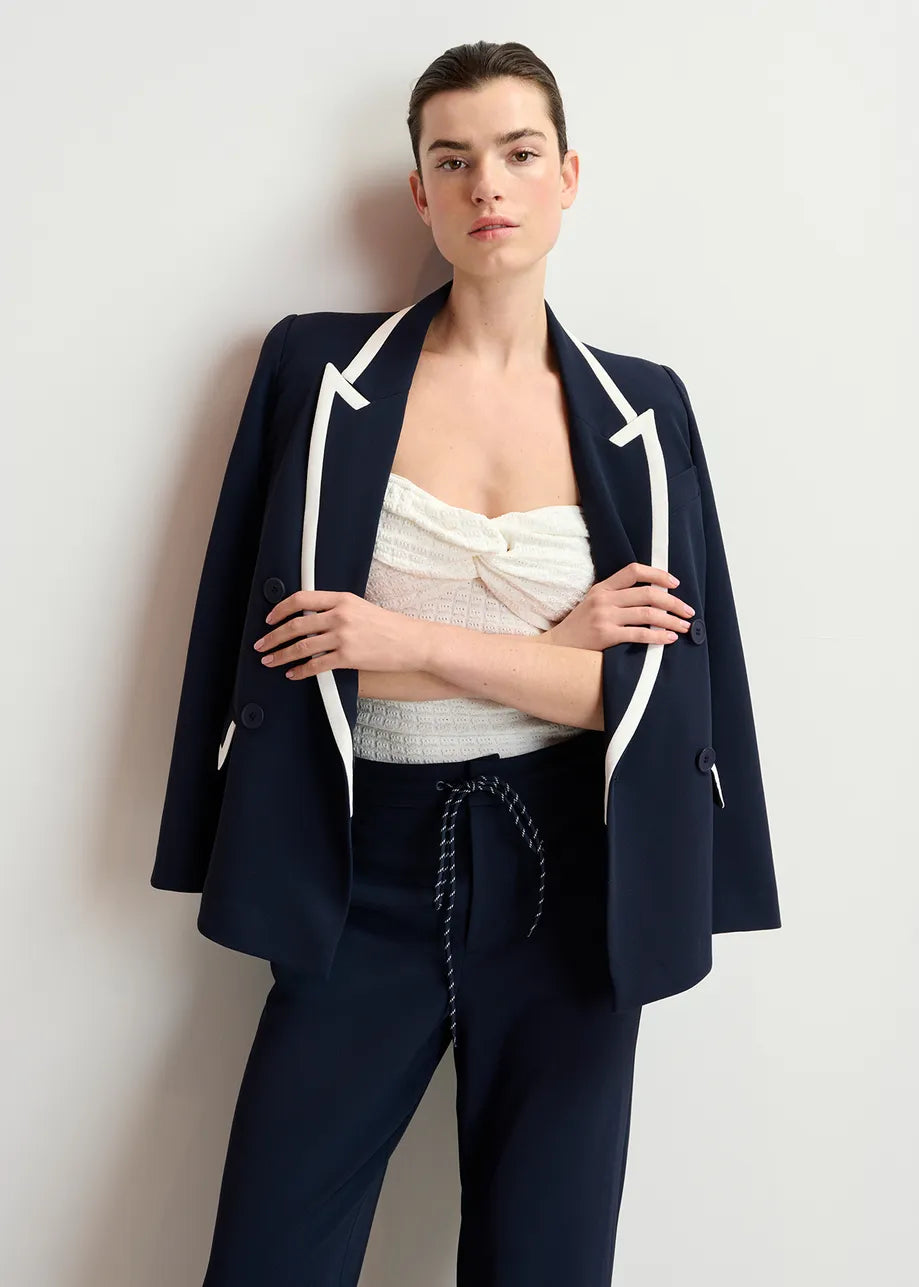 A person in a navy blazer with white trim, the Essentiel Antwerp Holo Knitted Strapless Top - White, and high-waisted navy pants stands against a light background, arms relaxed by their sides, looking forward.