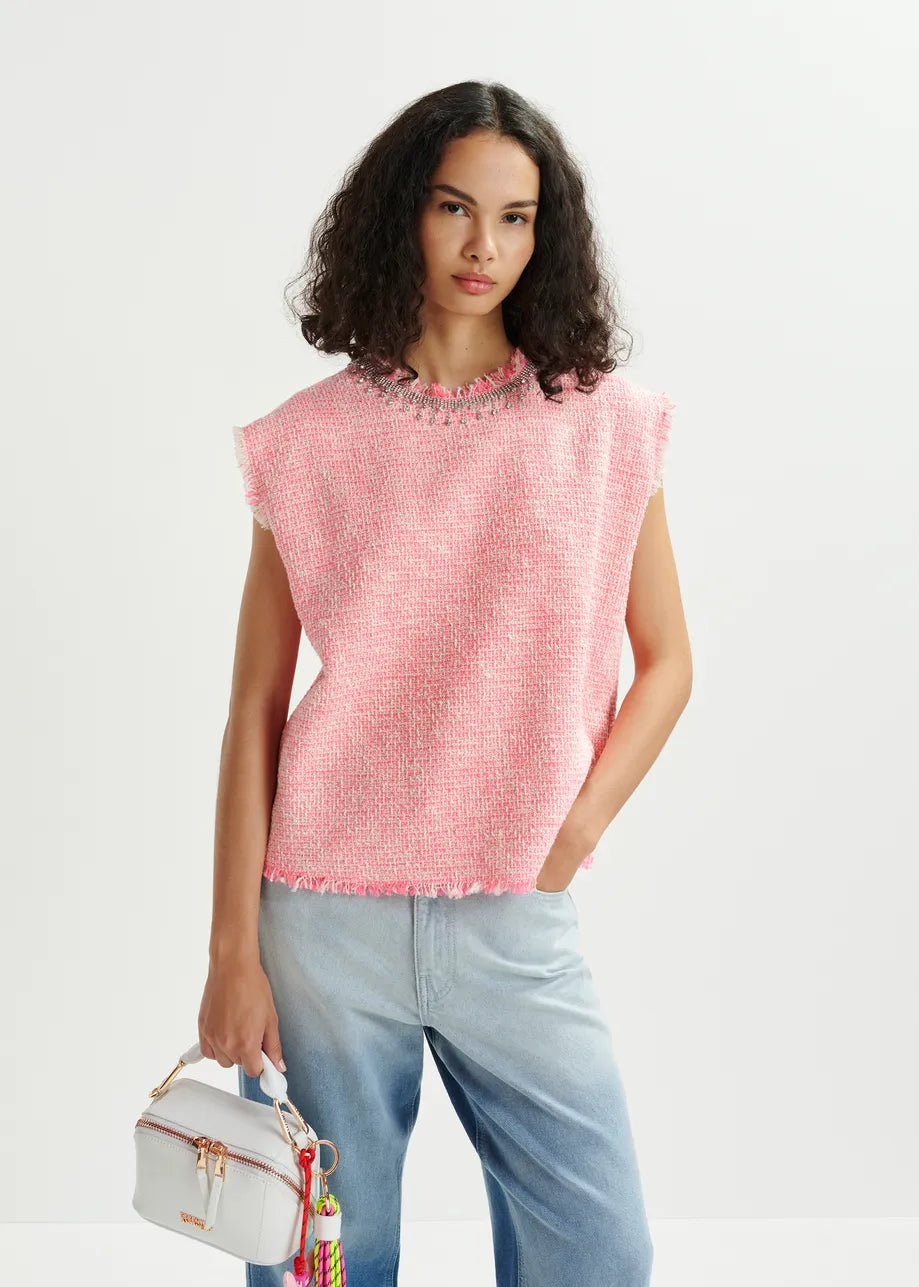 A person with curly hair wears the Essentiel Antwerp's Honduras Sleeveless Top in pink and light blue jeans, holding a small white handbag adorned with colorful tassels and subtle sequin embroidery.