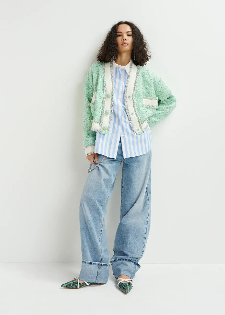 Someone wearing the Hoola Cropped Tweed Cardigan by Essentiel Antwerp in a green textured finish over a striped shirt, paired with wide-leg jeans and patterned shoes, stands against a plain white background.