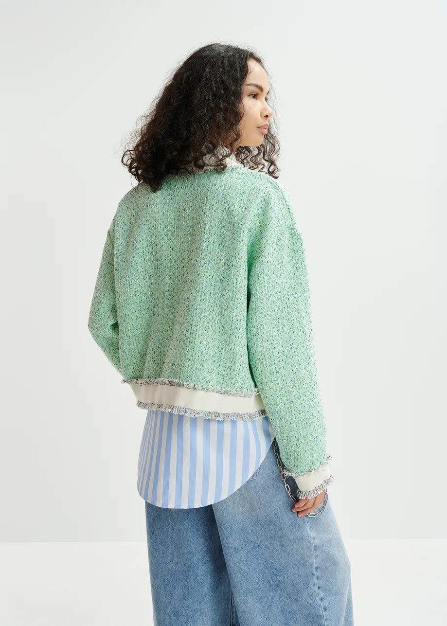 A person with curly hair wears Essentiel Antwerp's Hoola Cropped Tweed Cardigan, an oversized green textured jacket, along with a blue and white striped shirt and blue jeans, looking to the side against a white background.