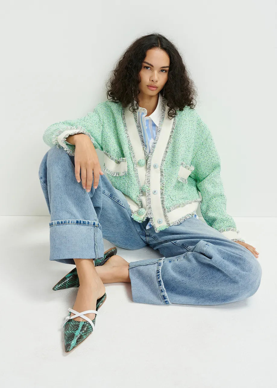 A person sits on the floor in an oversized Hoola Cropped Tweed Cardigan by Essentiel Antwerp, paired with blue jeans and pointed shoes, against a plain background.