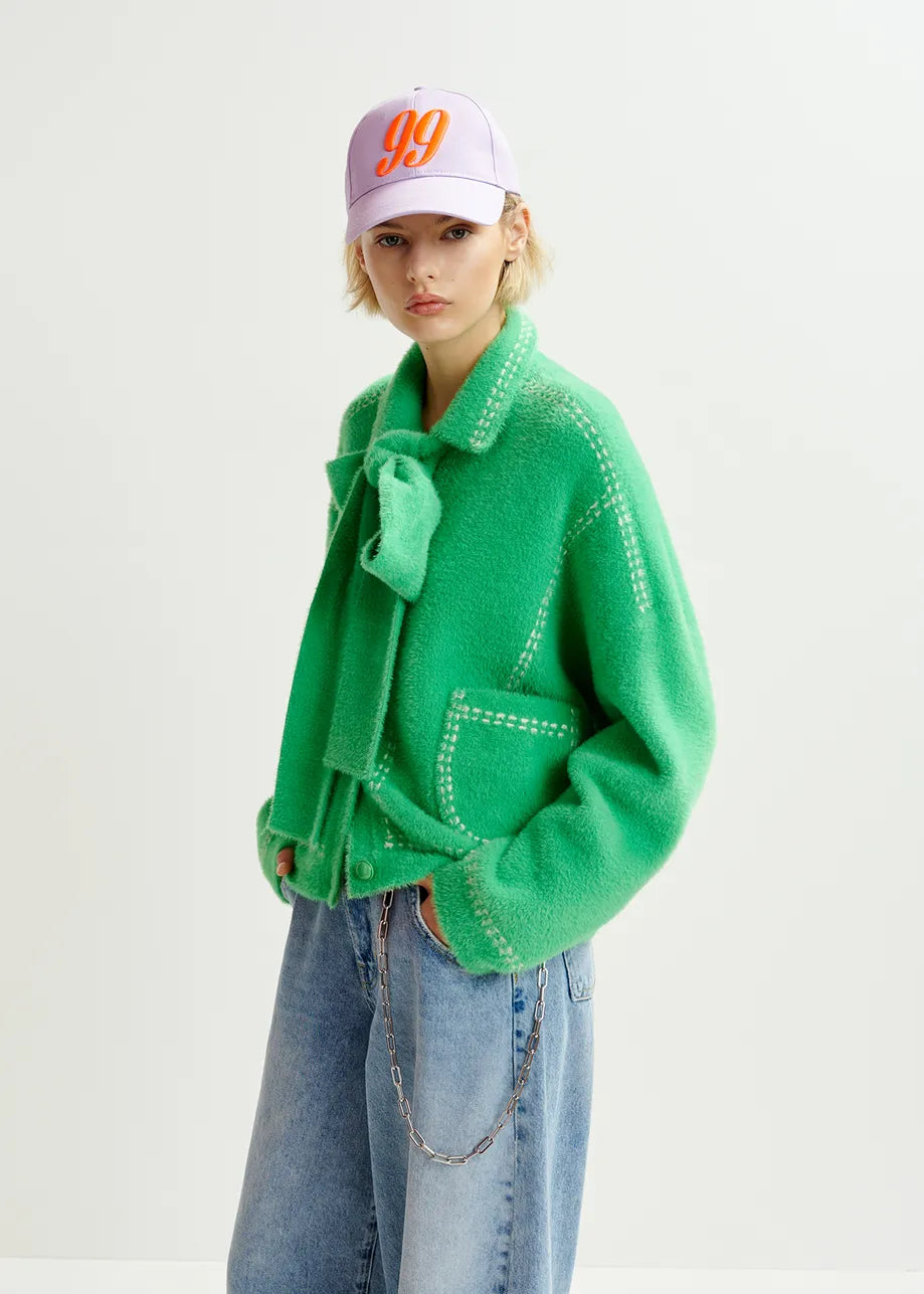 Clad in Essentiel Antwerp's Hoops Contrast-Knit Cardigan - Green, detailed with contrasting stitching and a bow, along with a light lavender "99" baseball cap and loose-fitting blue jeans featuring a chain, a person stands confidently against a plain backdrop.