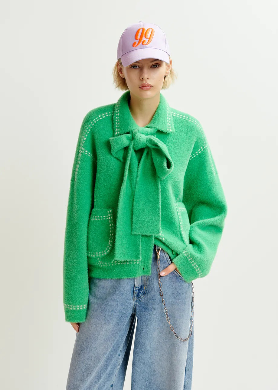 Someone flaunts an Essentiel Antwerp Hoops Contrast-Knit Cardigan in green, styled with wide-legged jeans and a pink cap featuring the number "99.