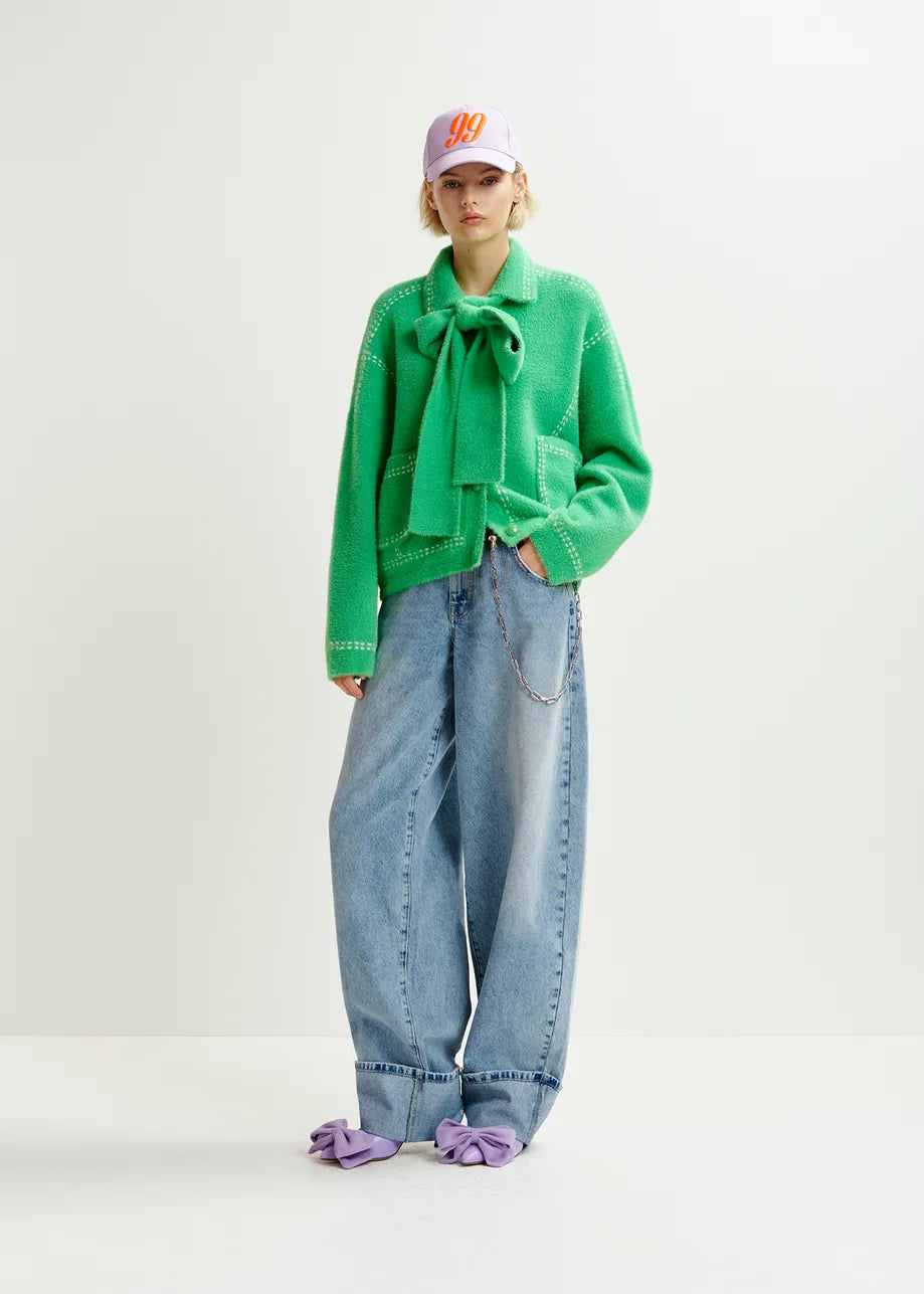 A person in a stunning Essentiel Antwerp Hoops Contrast-Knit Cardigan - Green, with wide-leg jeans, purple bow shoes, and a white cap stands against a plain background.