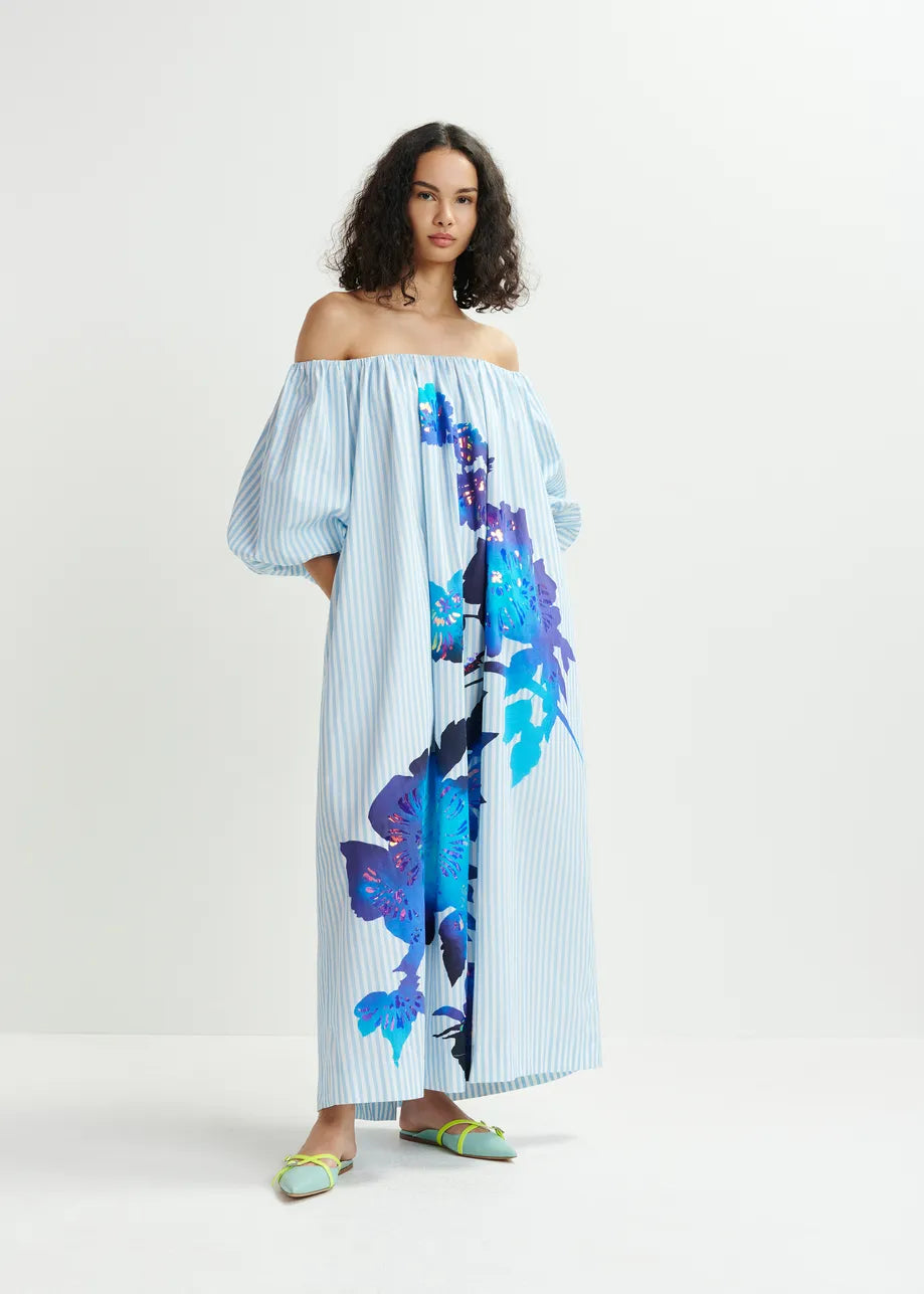 A woman stands gracefully in the Horpio Off-Shoulder Dress by Essentiel Antwerp, crafted from 100% cotton with light blue stripes and a floral foil print on a plain light background, exuding effortless elegance.