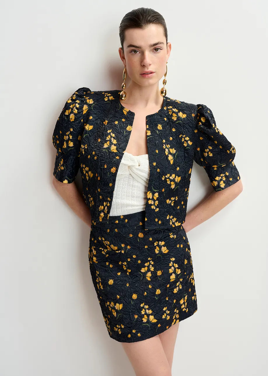 Dressed in an Essentiel Antwerp Hosha Cropped Jacket - Black with yellow floral patterns, the person pairs it with a matching skirt and large gold earrings. The oversized fit provides a modern twist, embracing sustainability with recycled polyester, all against a plain background.