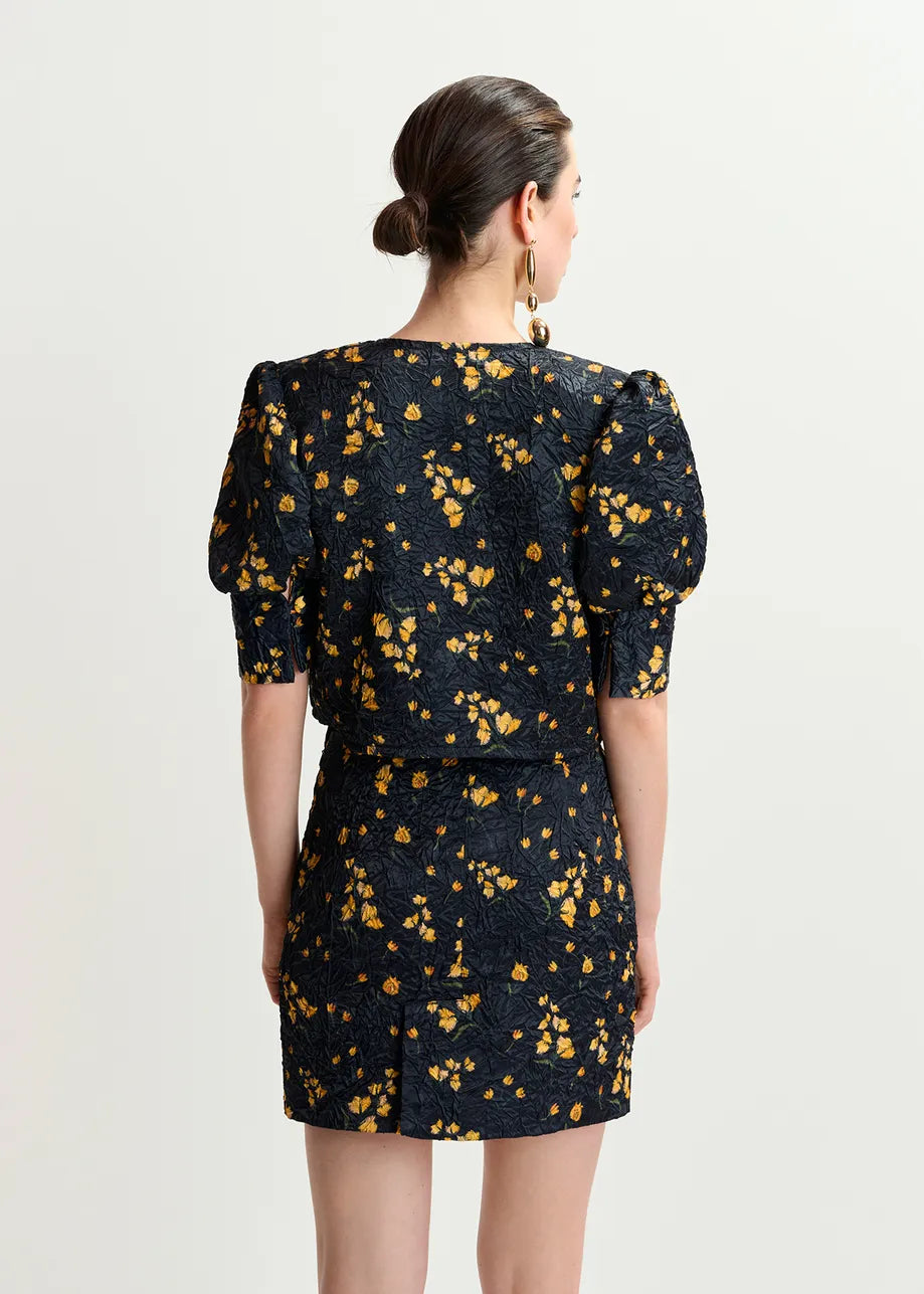 A woman faces away in a Hosha Cropped Jacket by Essentiel Antwerp. The black jacket has yellow floral patterns and puffed sleeves, offering an oversized fit for a modern touch. Her bun hairstyle and gold hoop earrings complete the look against a white background, embracing eco-conscious fashion with recycled polyester.