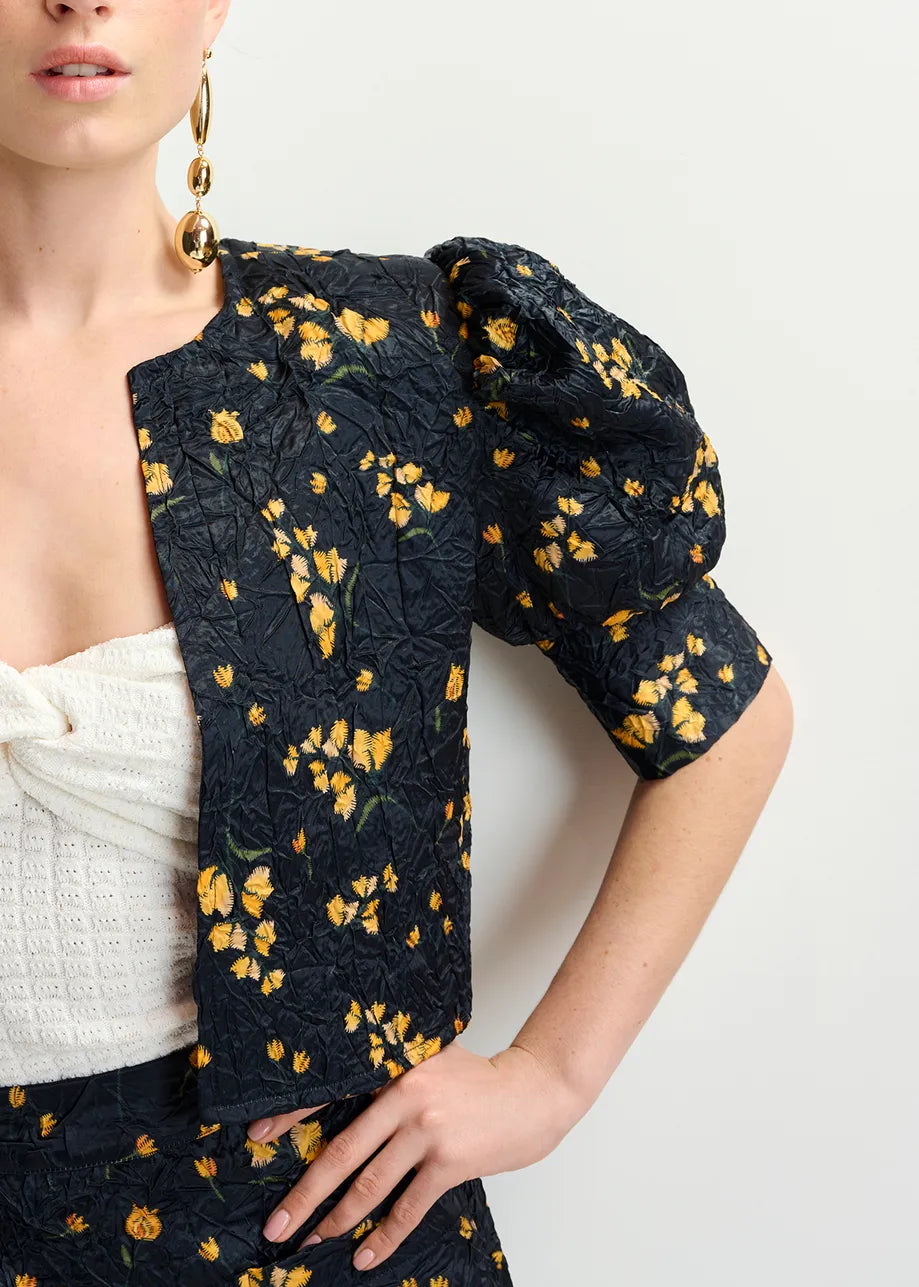 A person exudes confidence in Essentiel Antwerp's Hosha Cropped Jacket in black with yellow florals, paired with a white textured top and large gold earrings. Their right hand rests on their hip, nodding subtly to sustainability with the jacket's recycled polyester fabric.