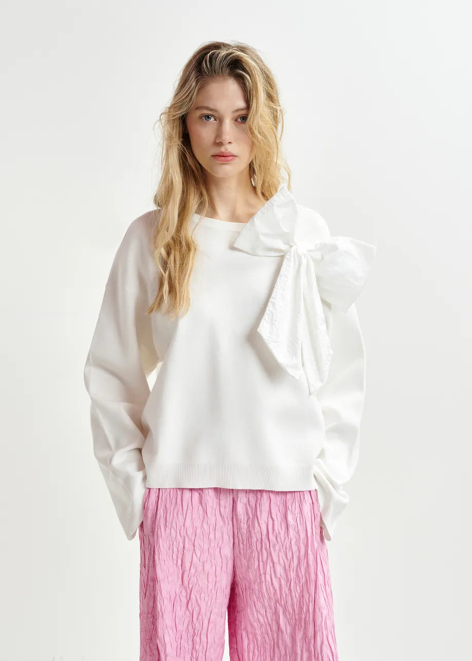 A person dressed in a white soft fine-knit "Hoslo Sweater Removable Bow" by Essentiel Antwerp and pink crinkled trousers stands against a plain background.