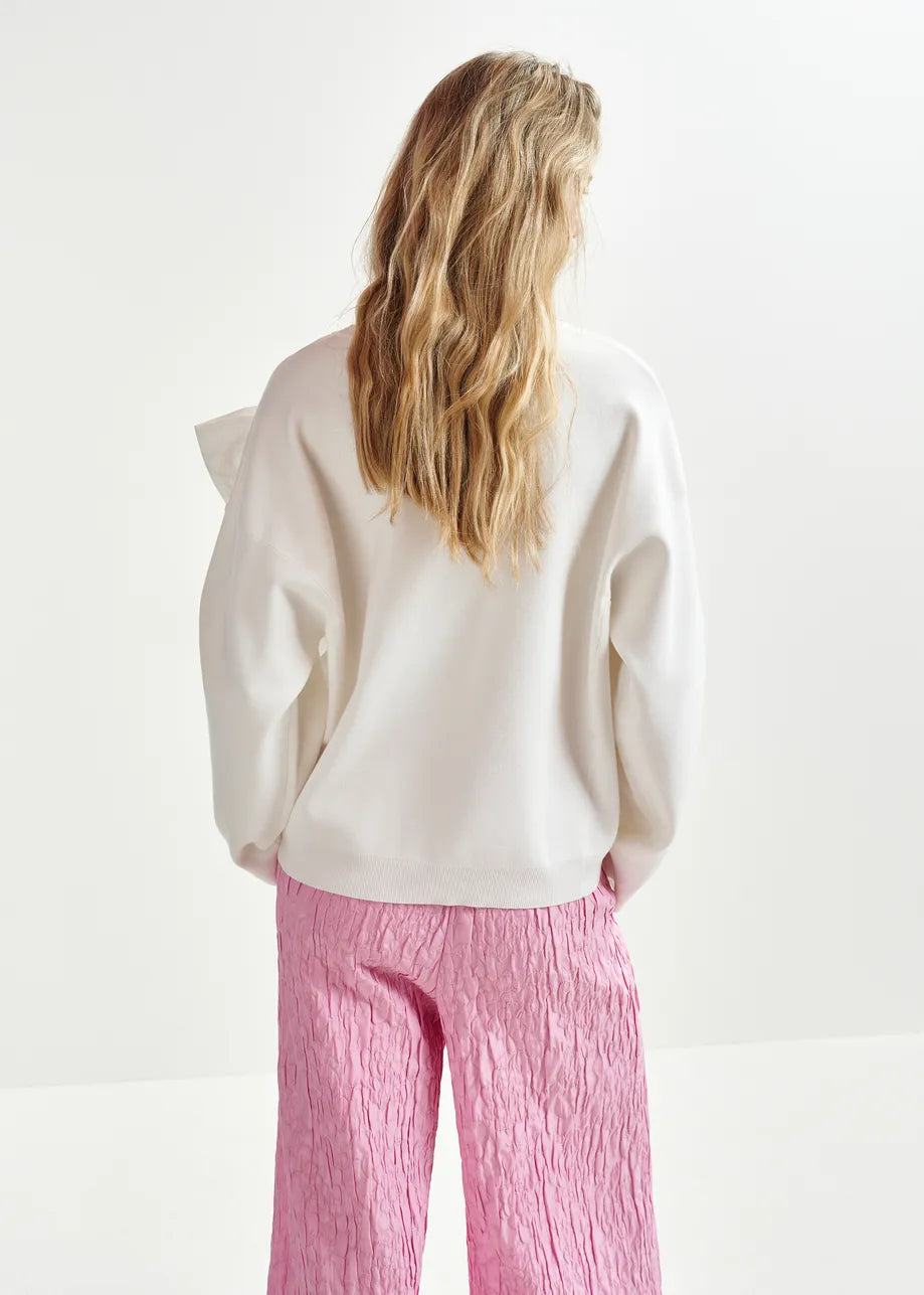 A person with long blonde hair wears an Essentiel Antwerp Hoslo Sweater with a removable bow in white and pink textured pants, standing with their back to the camera against a plain background.