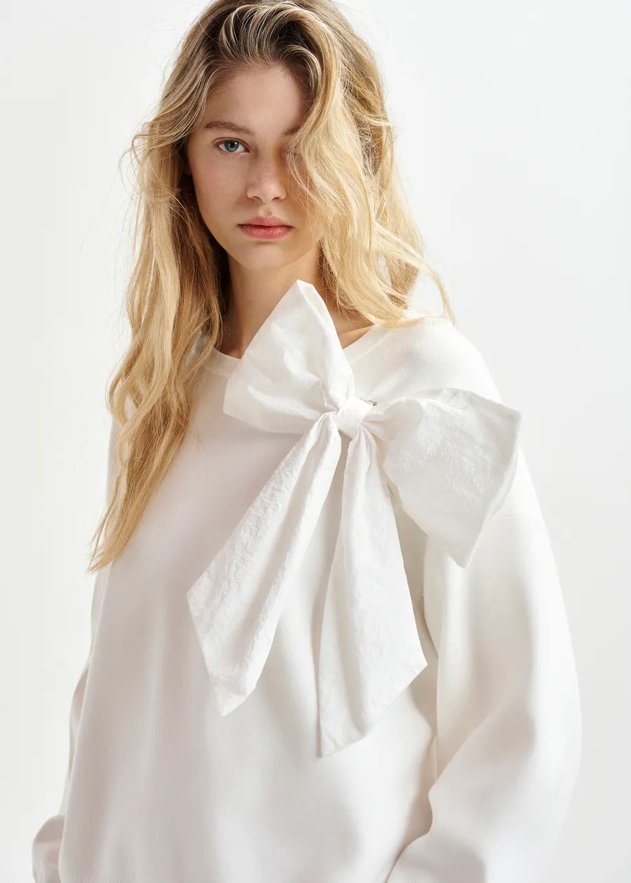 A person with long blonde hair wears the Hoslo Sweater Removable Bow in white by Essentiel Antwerp, which features a large removable bow on the shoulder.