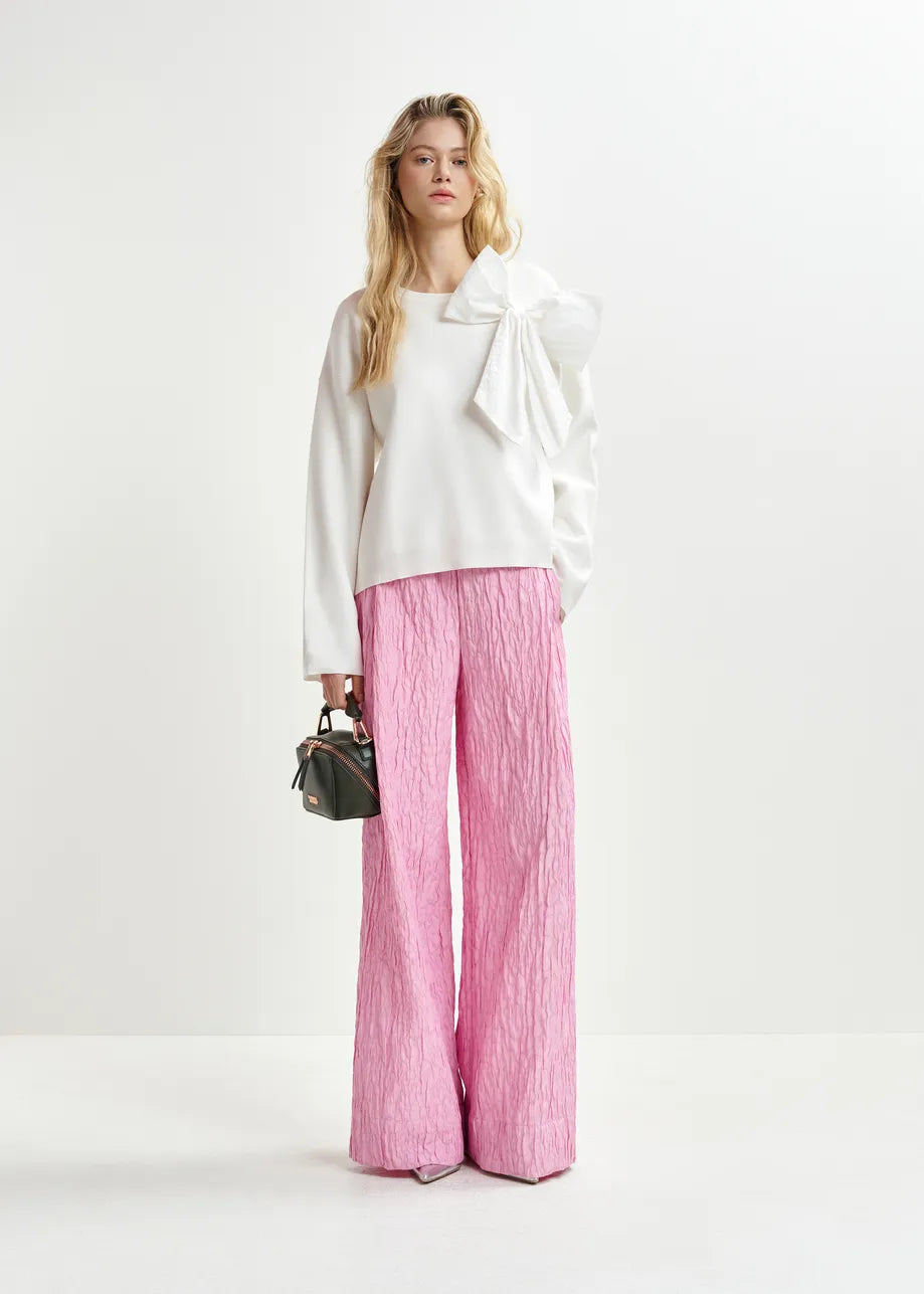 A person in a soft white Essentiel Antwerp Hoslo Sweater with a removable bow and pink wide-leg pants holds a small black handbag, standing against a plain background.