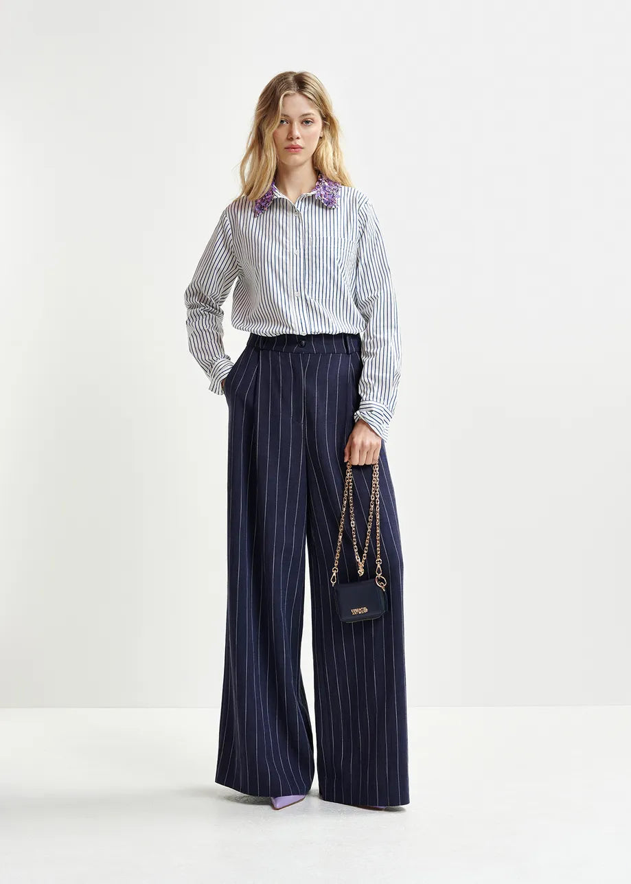 A woman dons Essentiel Antwerp's Hotel Pinstripe Trousers in navy, made from a recycled viscose blend, alongside a striped shirt and a small handbag with chain strap.