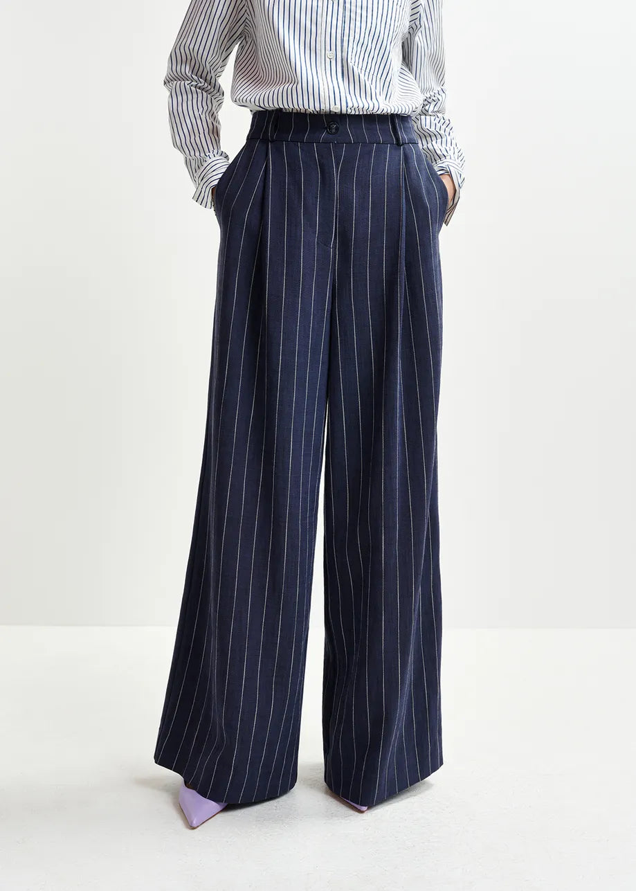 Dressed in Essentiel Antwerp's Hotel Pinstripe Trousers in navy, made from a recycled viscose blend, someone stands casually with hands in pockets, sporting a matching striped shirt.