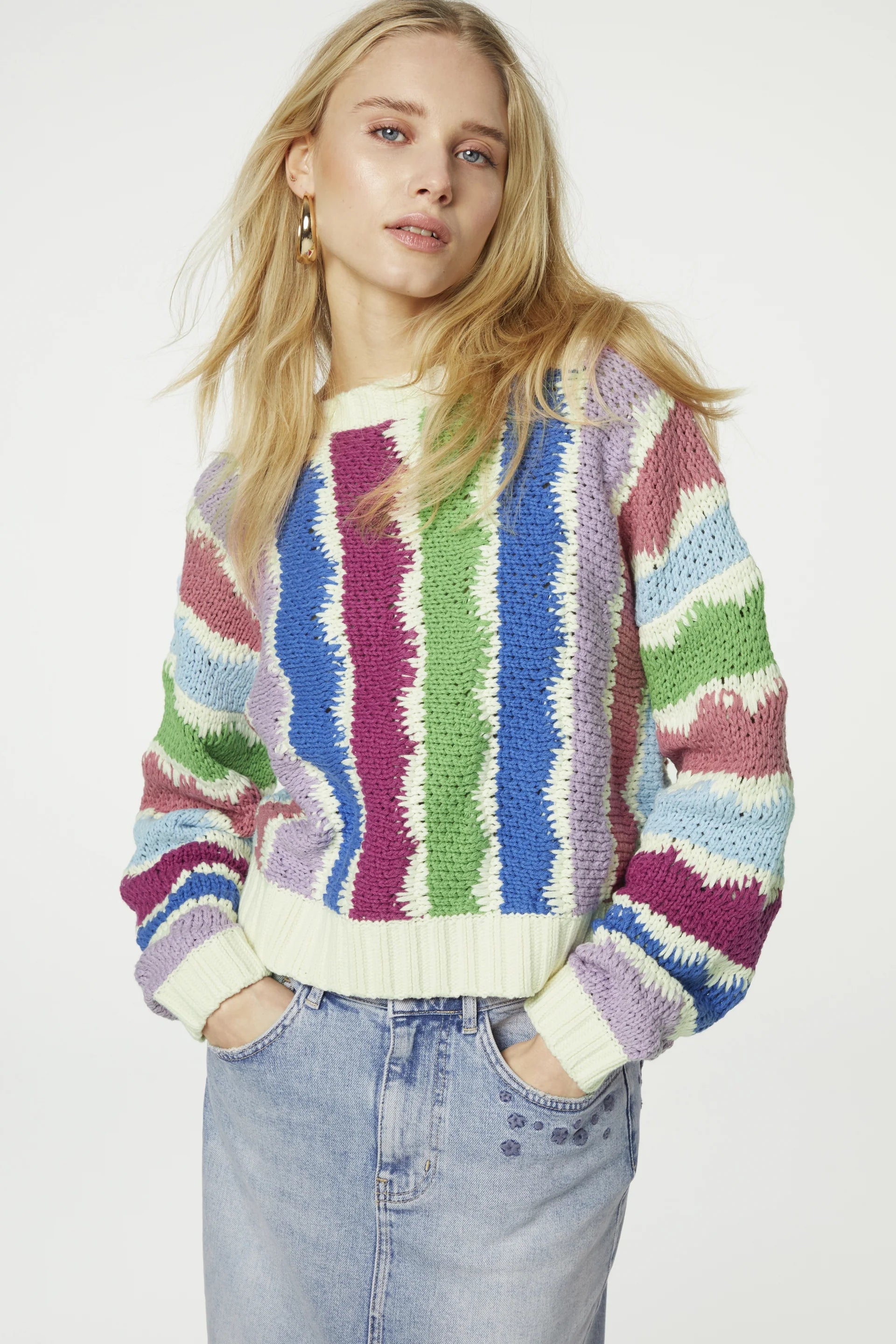 A person with long blonde hair wearing a Fabienne Chapot Heath Pullover - Multicolour and light blue jeans stands with hands in pockets against a white background.