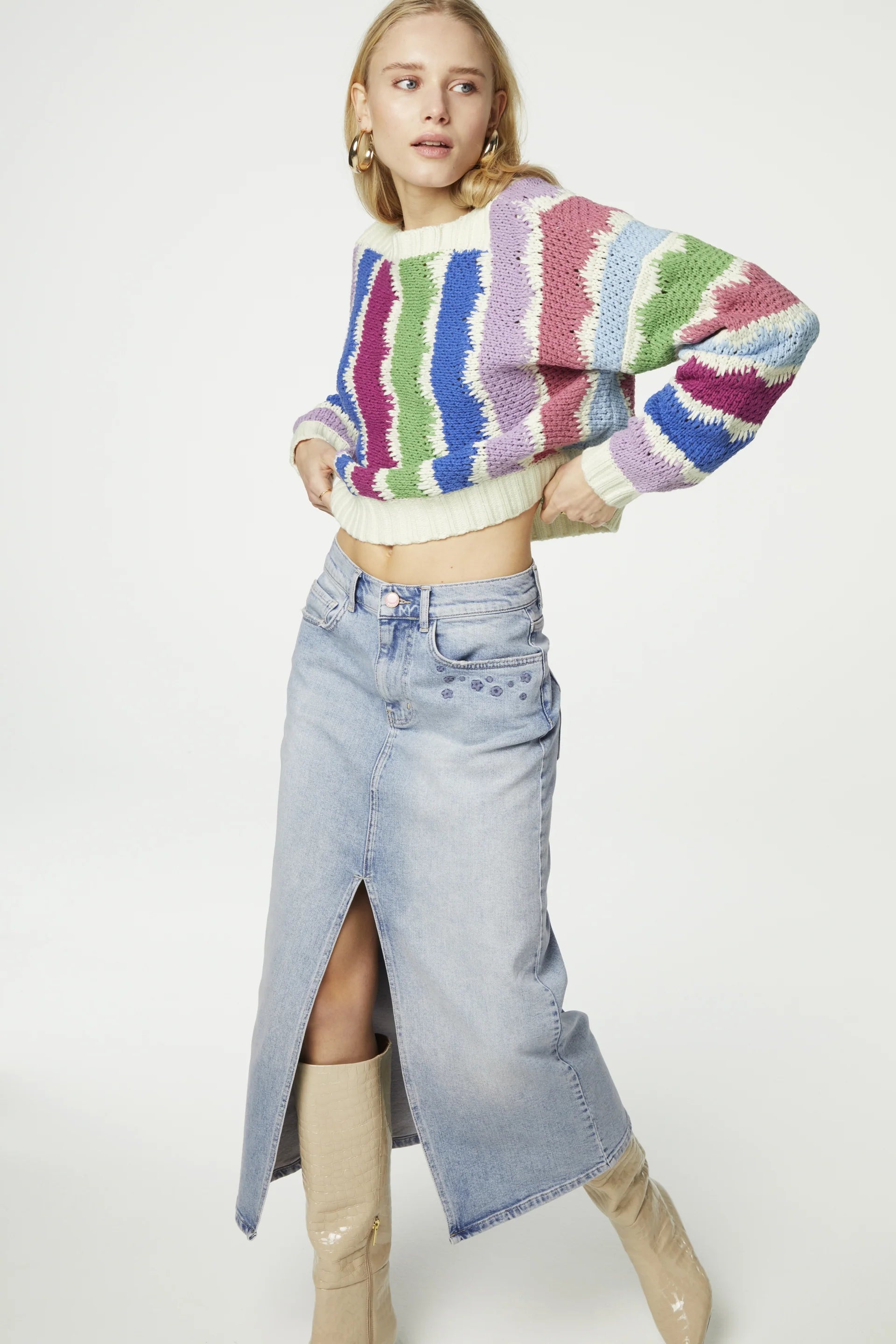 A woman wearing a Fabienne Chapot Heath Pullover - Multicolour with a light blue denim skirt that features a front slit stands against a white background. She has blonde hair and is looking to the side.