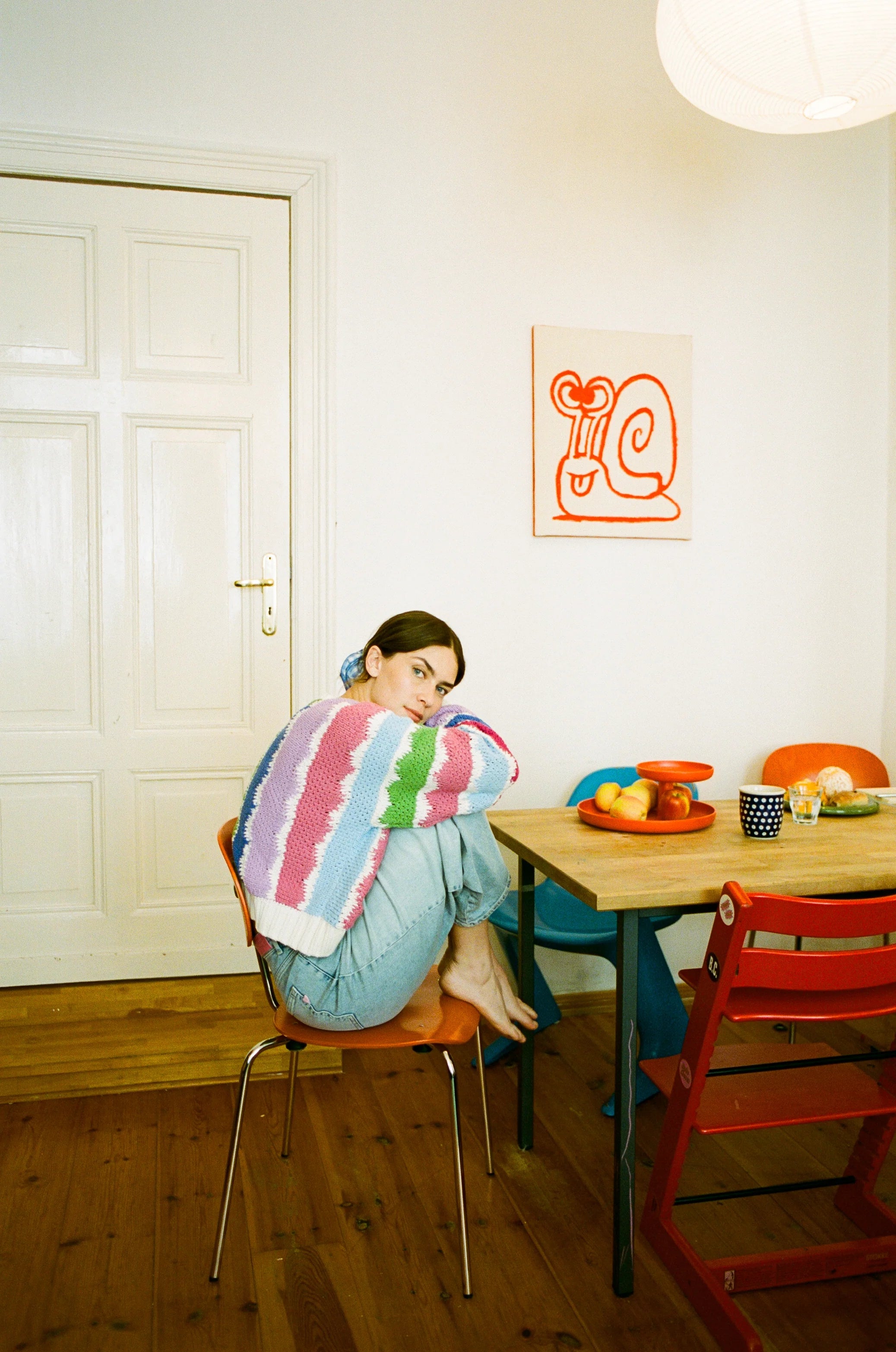 A person sits on a chair in a cozy room near a wooden table with a red chair, wearing the Heath Pullover - Multicolour by Fabienne Chapot and relaxed fit jeans, with food items on the table and an orange art piece on the wall.