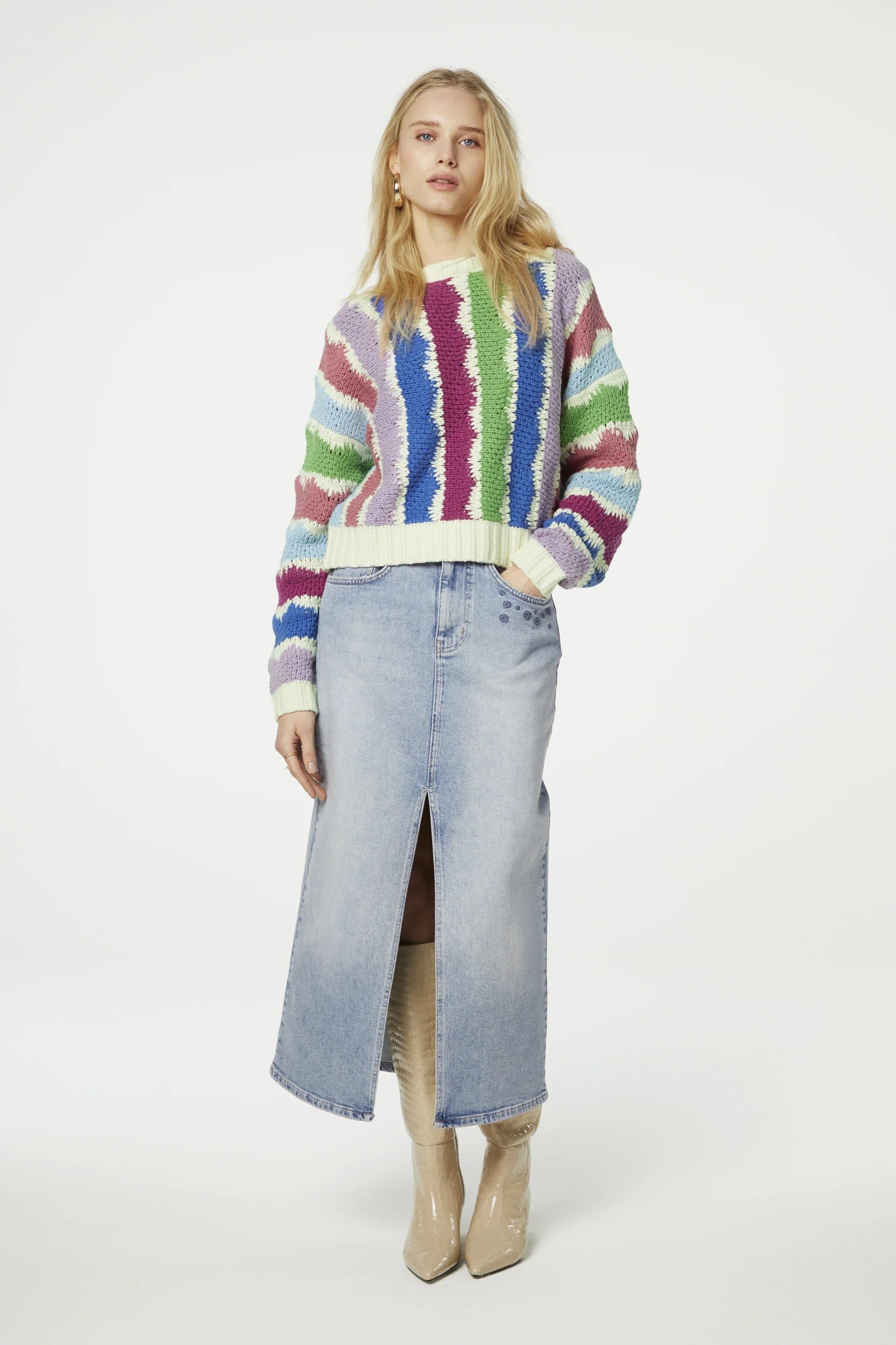 A blonde woman wearing a relaxed-fit Fabienne Chapot Heath Pullover in multicolour and a long denim skirt with a front slit stands against a white background. She is also wearing knee-high beige boots.