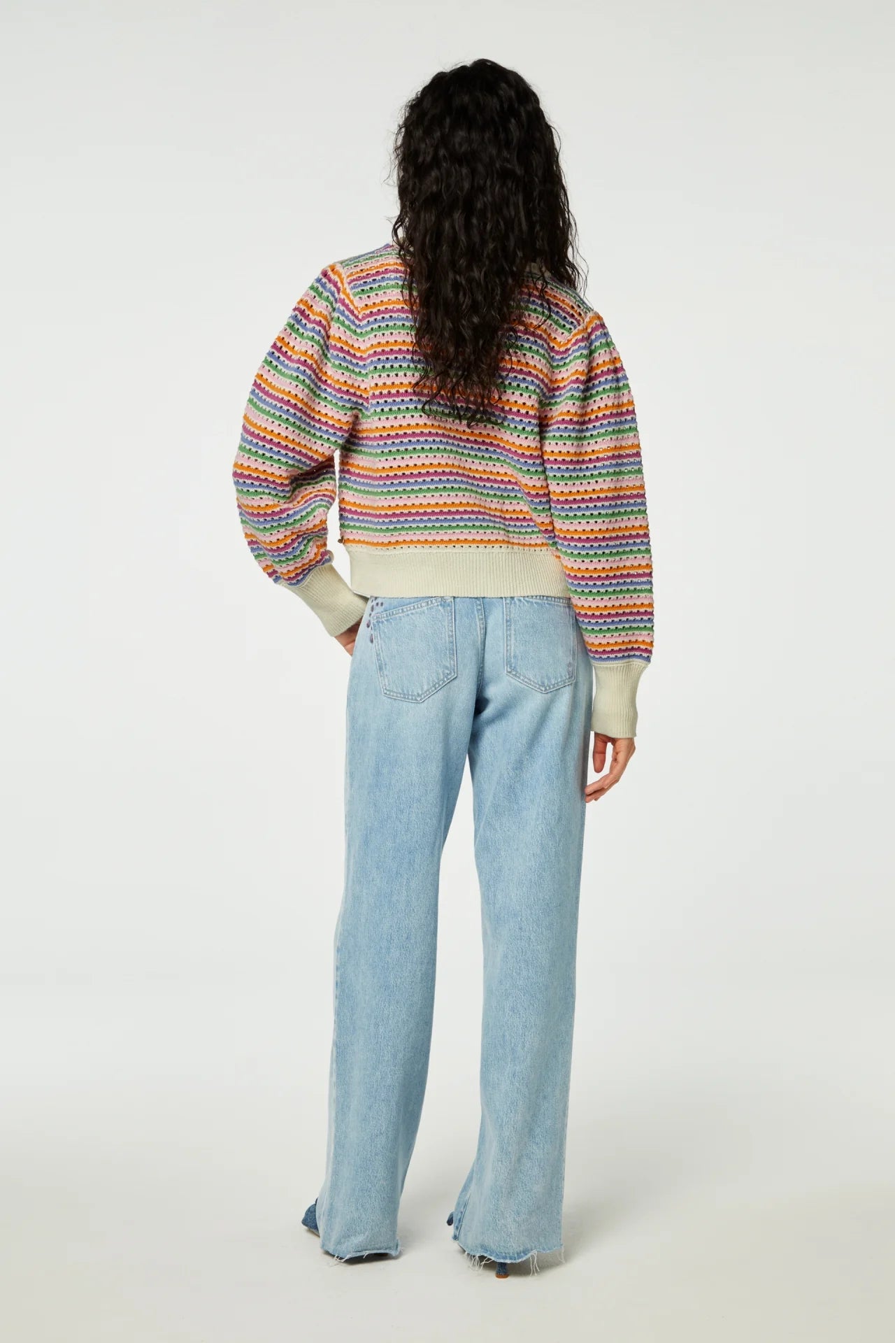 Woman from behind wearing a colorful striped Fabienne Chapot Heather Cardigan with balloon sleeves and blue jeans against a plain background.