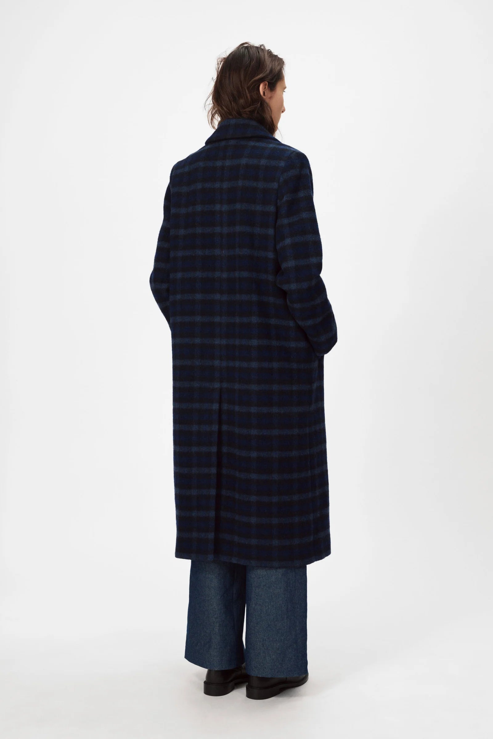 A person stands with their back to the camera, wearing the Nué Notes Helsinki Coat in navy and wide-leg jeans, set against a plain white background.