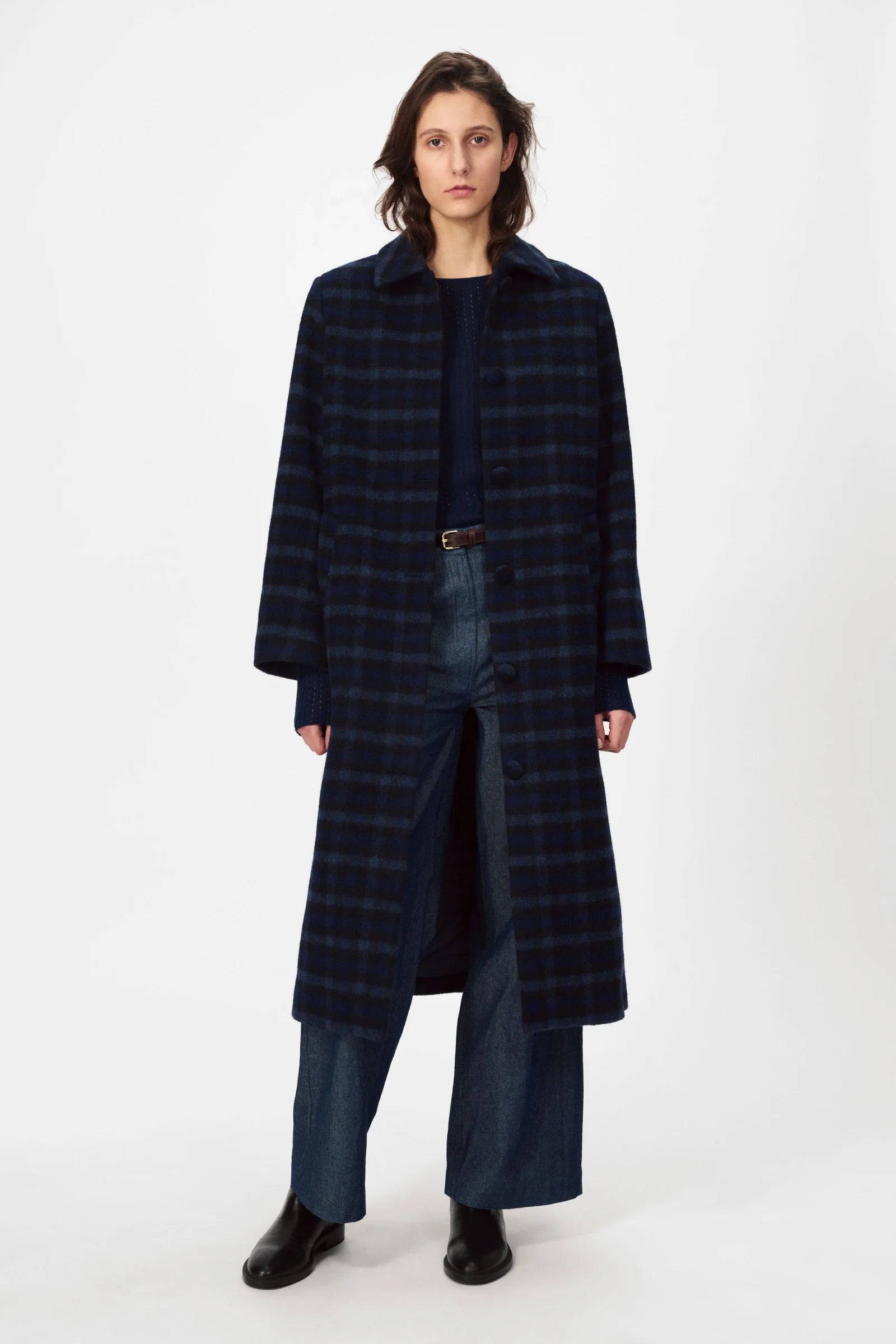 A person stands against a white background wearing the Nué Notes Helsinki Coat in navy, paired with wide-leg jeans, black boots, and a dark top.