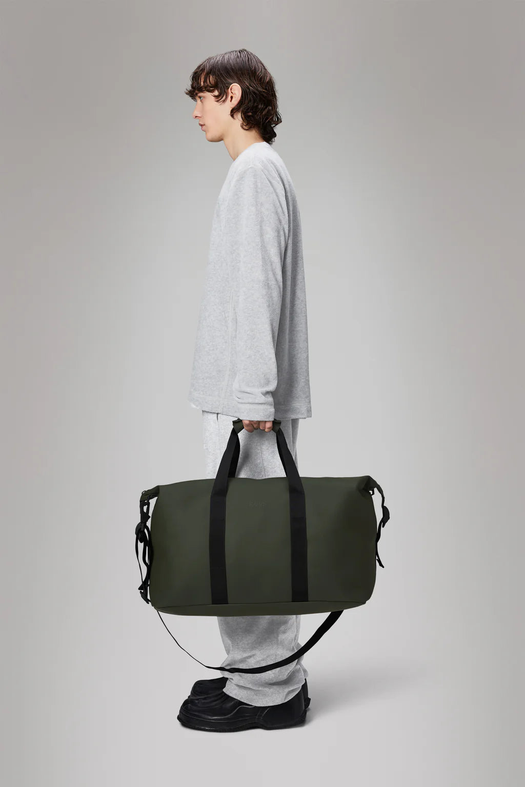 A person in gray long-sleeve and pants stands sideways, holding a large green Hilo Weekend Bag crafted by Rains.