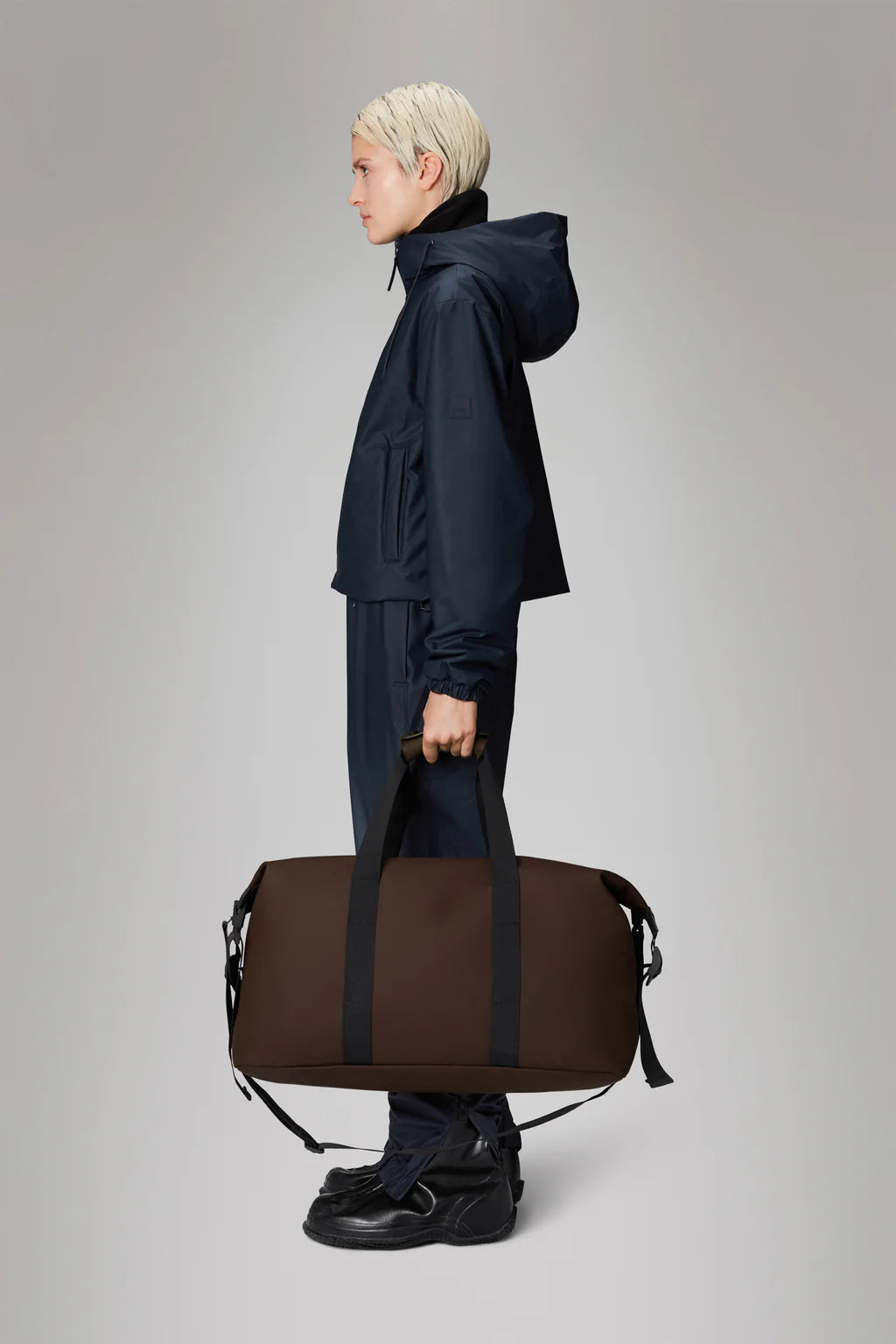 A person in a dark outfit stands against a gray background, holding the Hilo Weekend Bag - Frame by Rains, expertly crafted with their signature PU fabric.