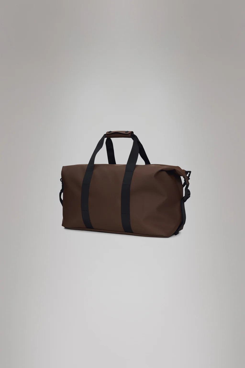 The Hilo Weekend Bag - Frame by Rains is designed in a brown duffel style, featuring black handles and a shoulder strap, crafted from their signature waterproof PU fabric. It impressively contrasts against the gray background, making it perfect for an overnight getaway.