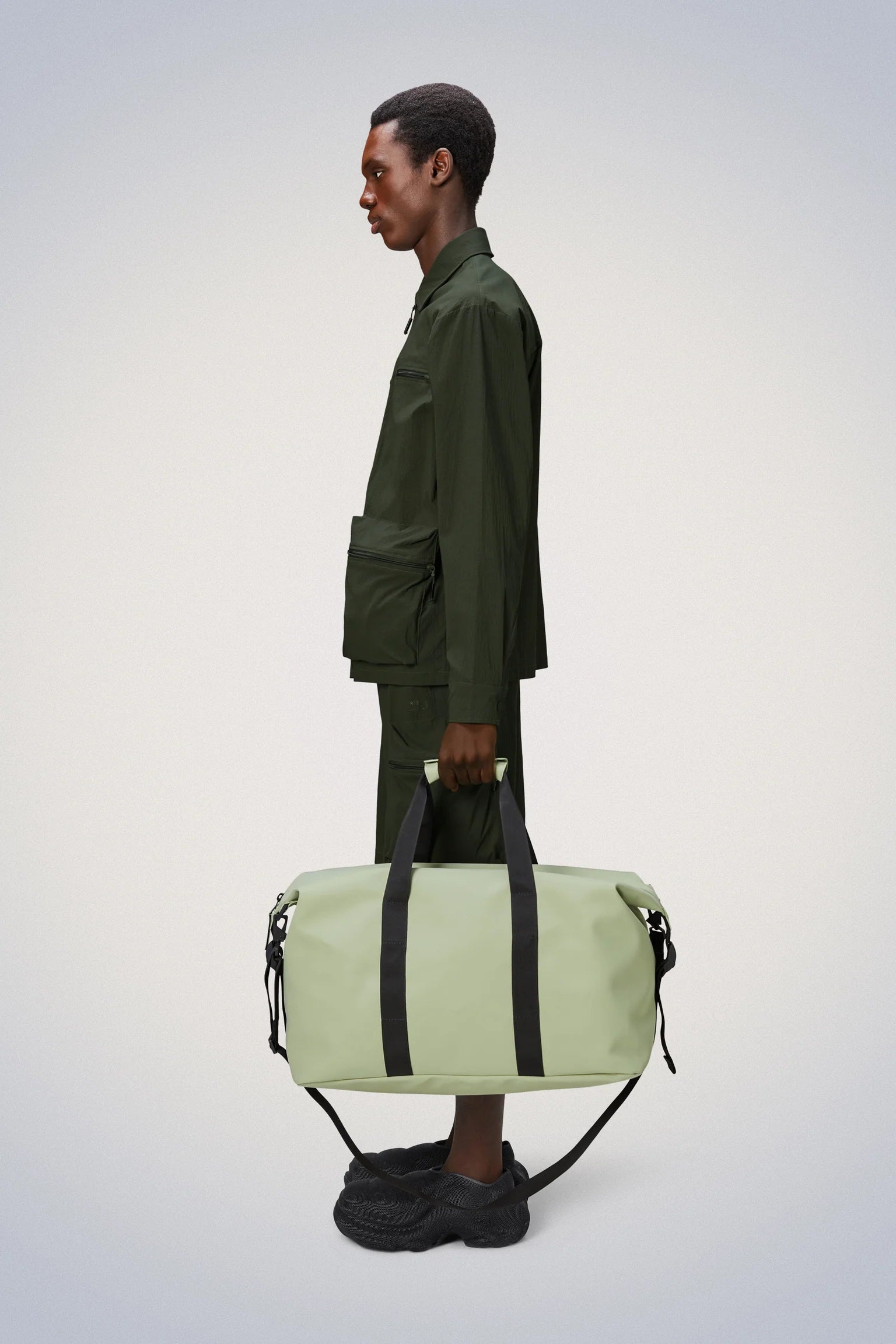 A man is holding a Rains Hilo Weekend Bag, his travel-favourite overnight bag, which happens to be green in color.