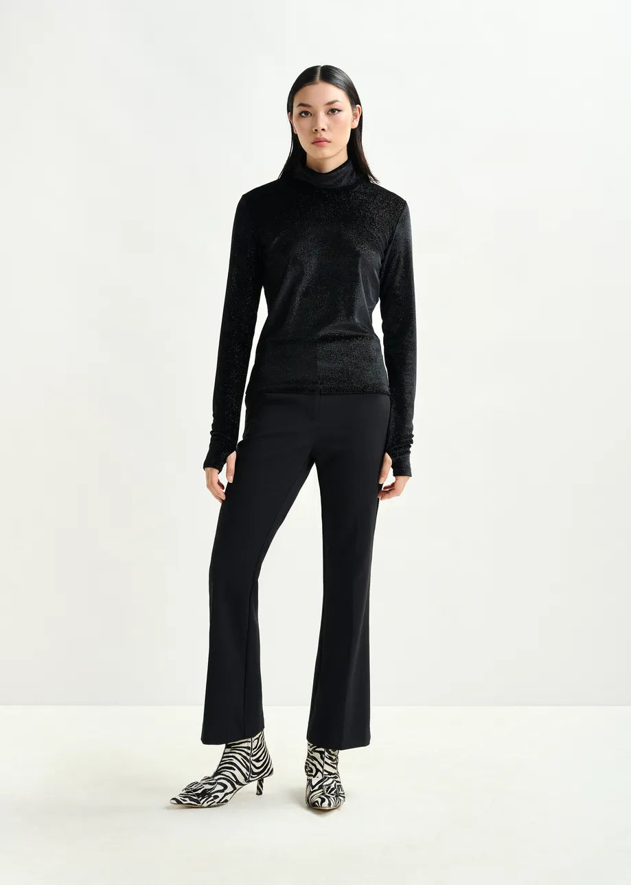 A woman stands against a plain background wearing a black turtleneck top, Essentiel Antwerp’s Ibell Flared Rhinestone Trousers in black, and zebra-print ankle boots.