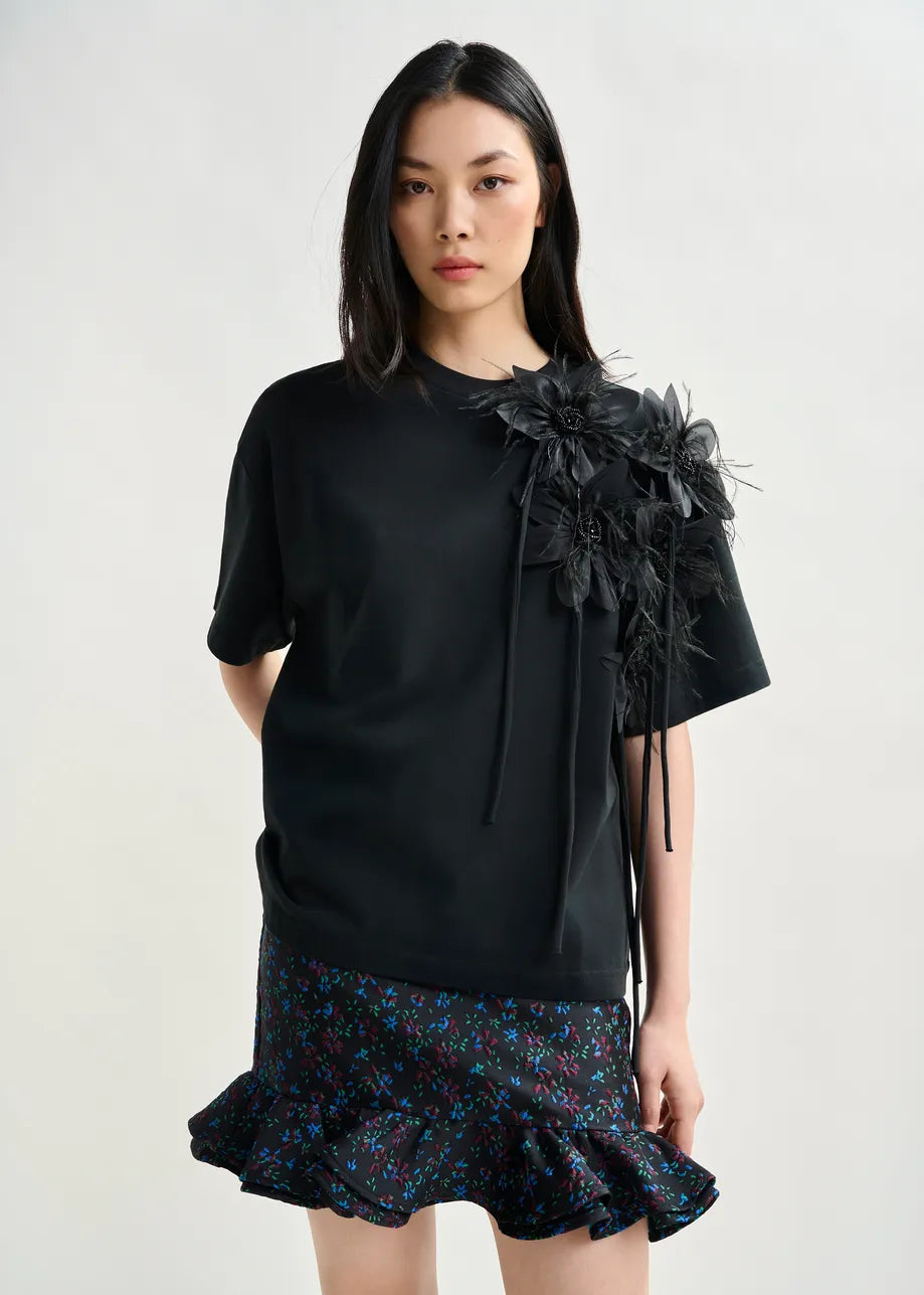 A woman stands against a plain background wearing the Essentiel Antwerp Ibiscus T-shirt in black, featuring floral embellishment on the shoulder, paired with a black skirt with blue and purple floral pattern and ruffled hem.