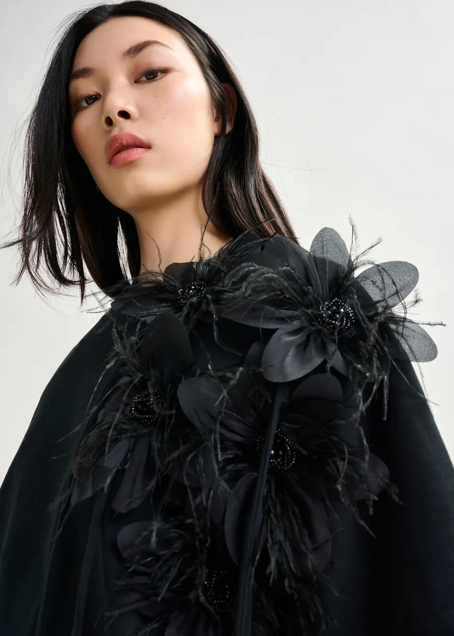 A woman models the Essentiel Antwerp Ibiscus T-shirt in black, a relaxed-fit top featuring bold black floral embellishments and feathers, standing against a plain light background.