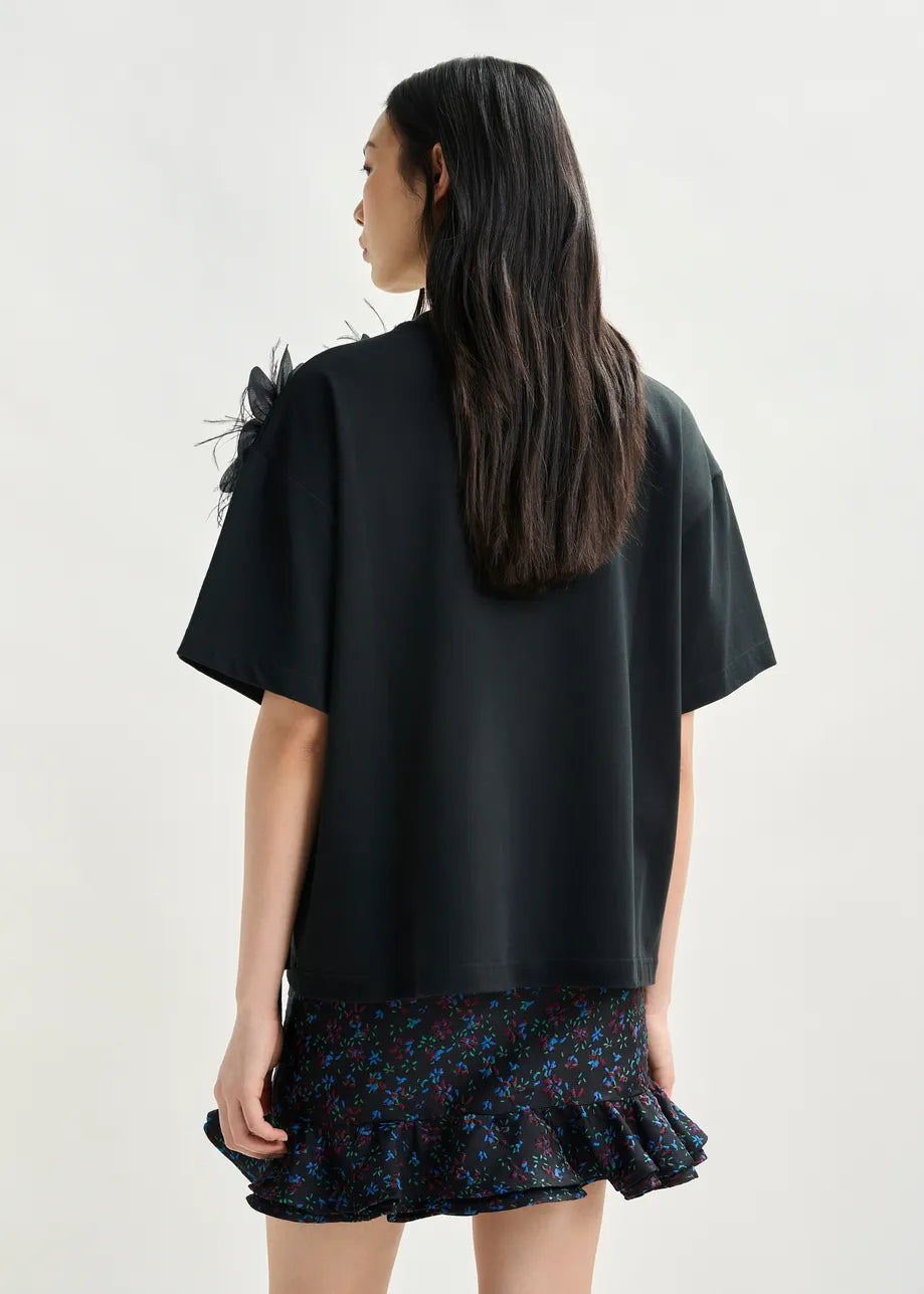 A woman with long dark hair, facing away from the camera, wears the Essentiel Antwerp Ibiscus T-shirt in black— a relaxed-fit 100% cotton top with a floral shoulder detail—paired with a dark ruffled mini skirt featuring a small floral print.