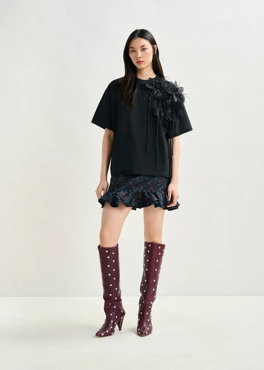 A woman stands against a plain background wearing the Ibiscus T-shirt in black by Essentiel Antwerp, paired with a ruffled mini skirt and knee-high maroon boots with white polka dots.