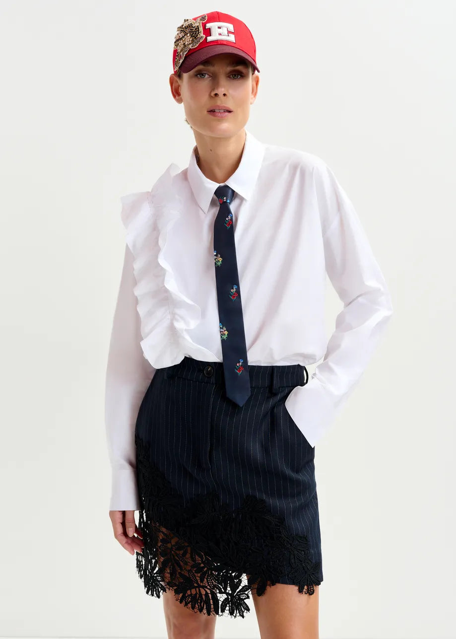 A person wears the Ibody Ruffle Shirt in white by Essentiel Antwerp, styled with a red cap, a dark pinstripe skirt with lace trim, and a navy floral tie.