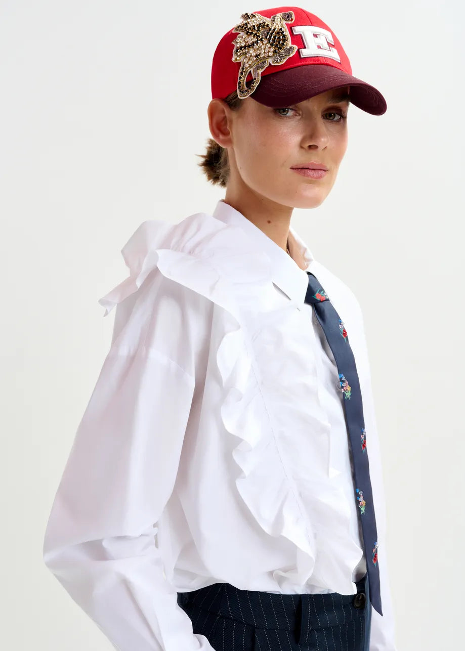 A person wearing the Essentiel Antwerp Ibody Ruffle Shirt in white, styled with a red embellished cap, dark floral tie, and pinstriped pants, poses against a plain background.