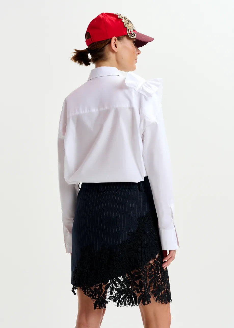 A person wearing the Essentiel Antwerp Ibody Ruffle Shirt in white, a black pinstripe skirt with lace trim, and a red baseball cap stands facing away from the camera.