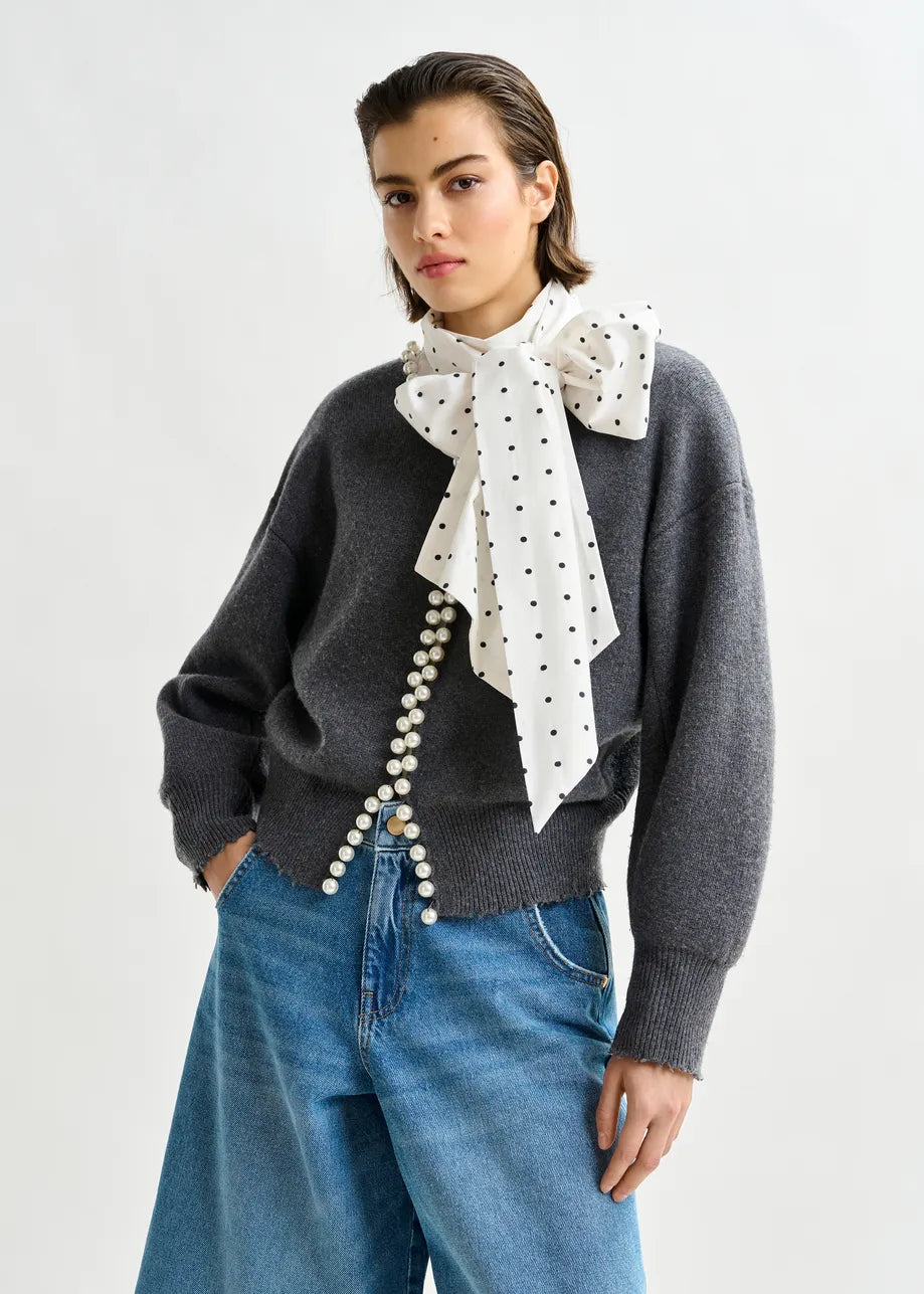 A woman poses against a plain background in the Essentiel Antwerp Ideas Pearl-Embellished Cardigan in grey, styled with a white polka dot bow scarf and relaxed-fit wide-leg blue jeans.