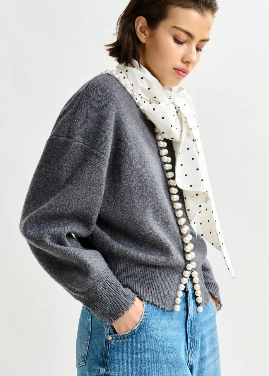 A woman wears the Essentiel Antwerp Ideas Pearl-Embellished Cardigan in grey, styled with blue jeans, a white polka dot scarf, and a string of pearls across her torso, standing against a plain background.