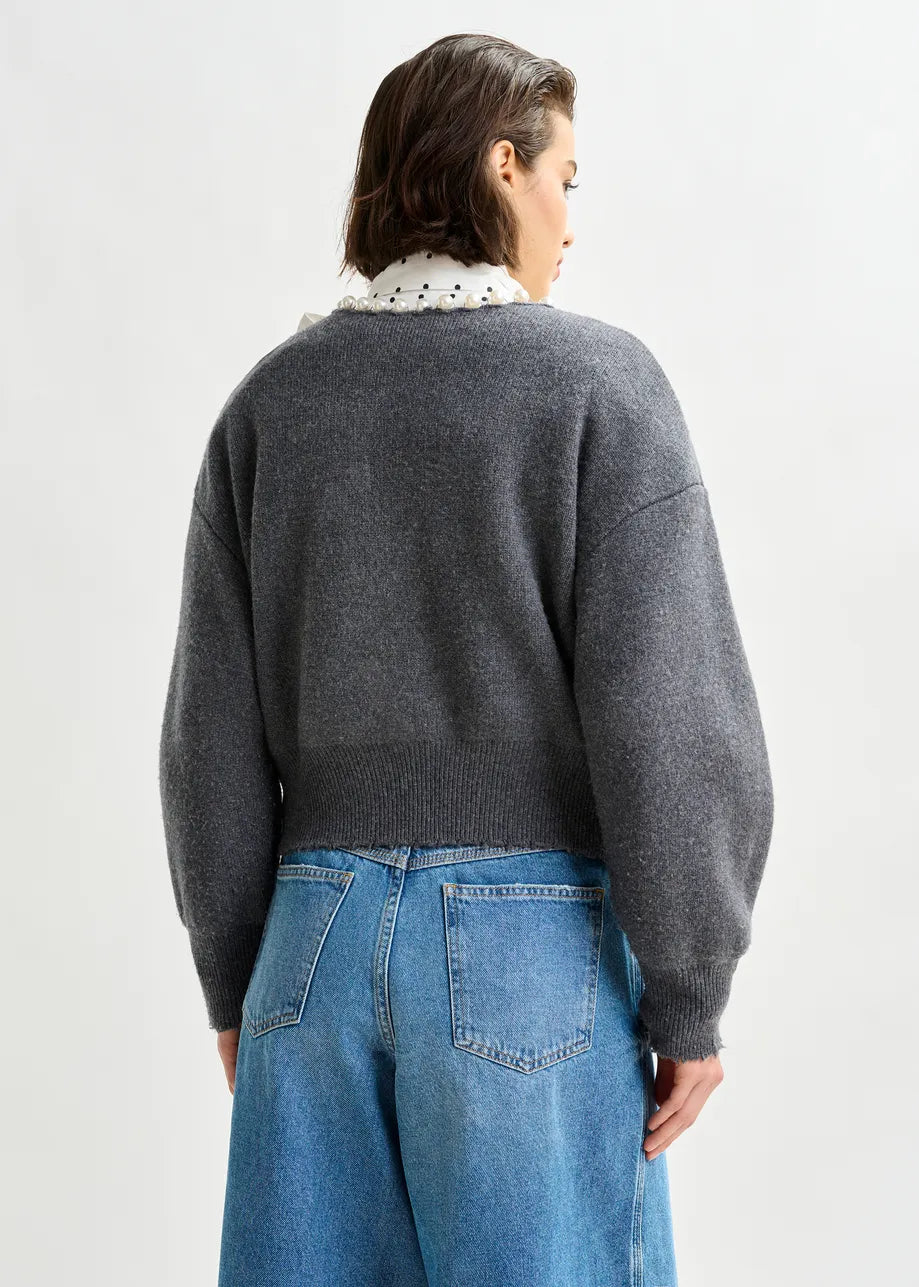 A person stands with their back to the camera, wearing the Essentiel Antwerp Ideas Pearl-Embellished Cardigan in grey over a white collared shirt and blue high-waisted jeans against a plain light background.