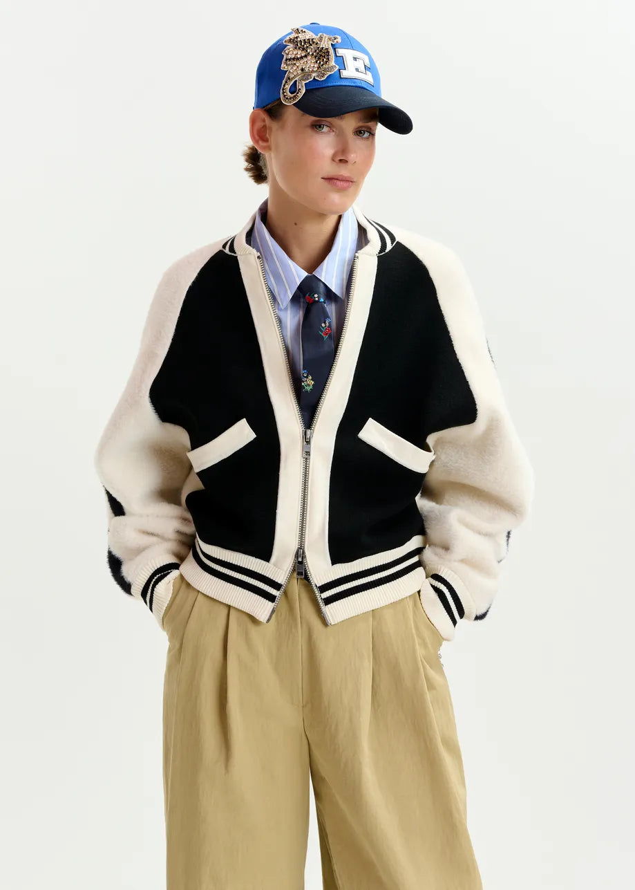 A person wears the Essentiel Antwerp Idol Knitted Bomber in black & white, paired with a blue embroidered cap, striped shirt with patterned tie, and beige pants against a plain background.