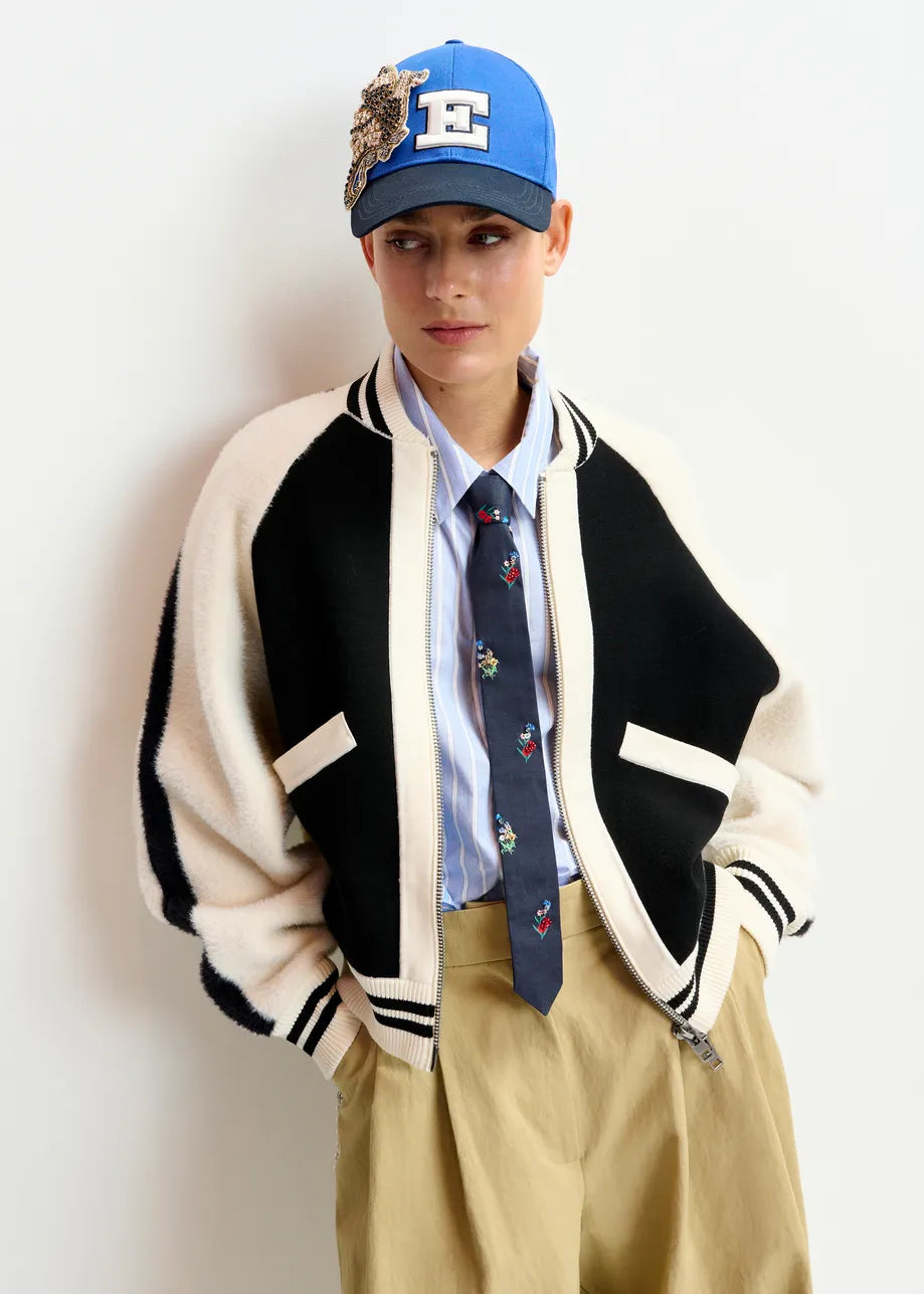 A person wears the Essentiel Antwerp Idol Knitted Bomber in black & white, paired with a blue embroidered tie, striped shirt, tan pants, and a blue embroidered patch cap, standing against a plain white background.