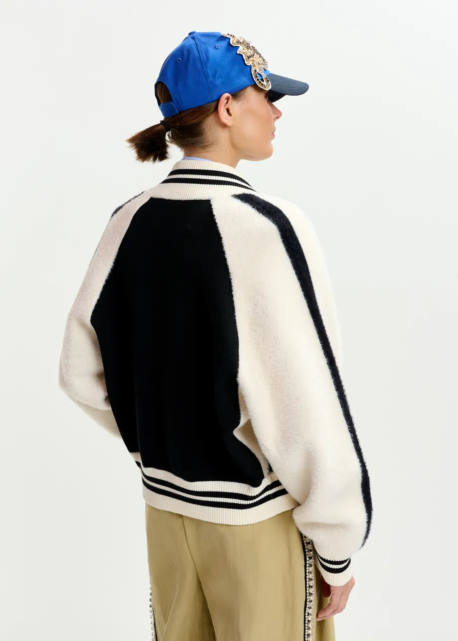 A person is seen from the back, wearing a blue cap, khaki pants, and the Essentiel Antwerp Idol Knitted Bomber in black & white, against a plain white background.