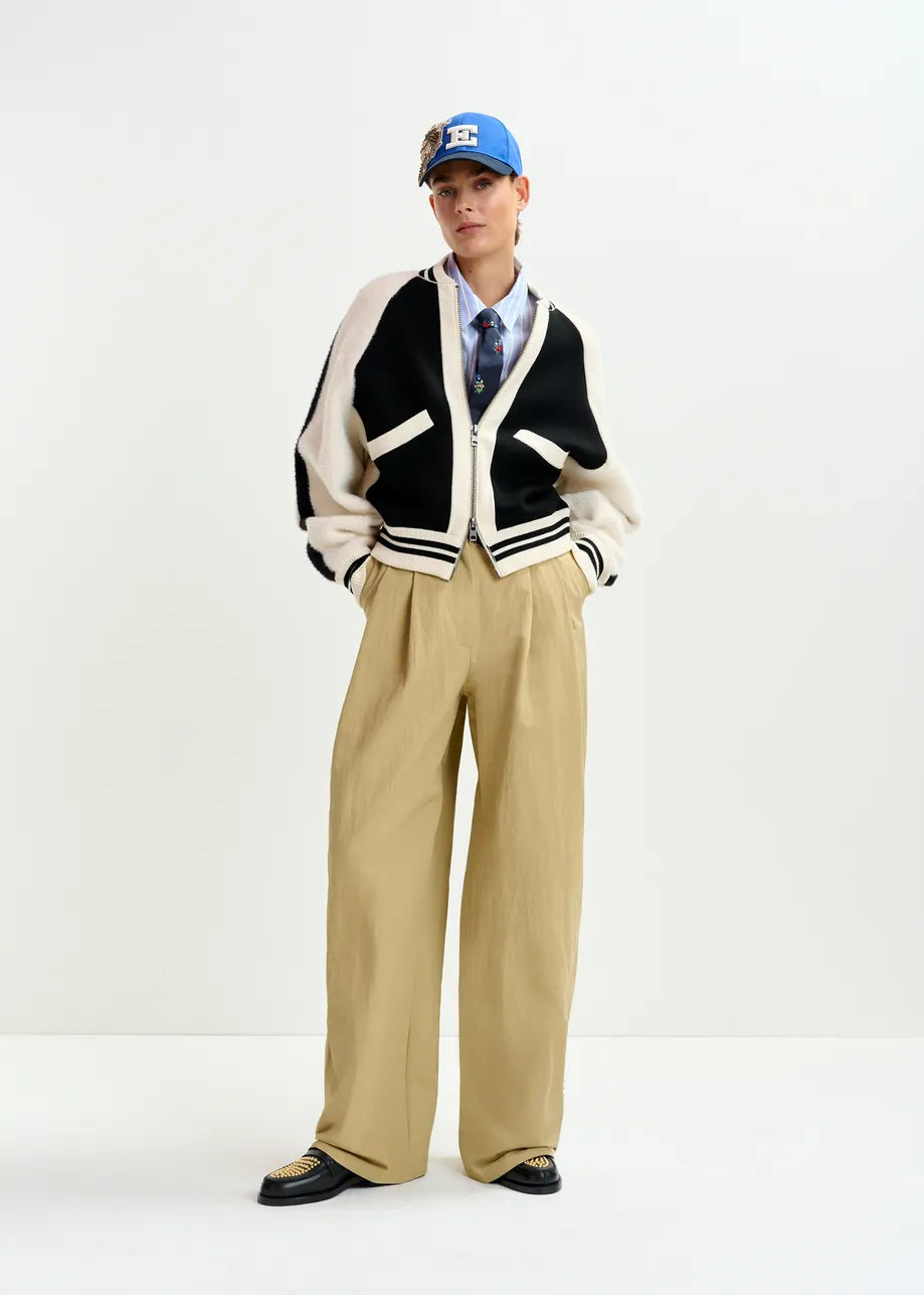 A person wears the Essentiel Antwerp Idol Knitted Bomber in black & white, paired with a blue cap, tie, light blue shirt, wide beige pants, and black shoes, standing against a plain white background.